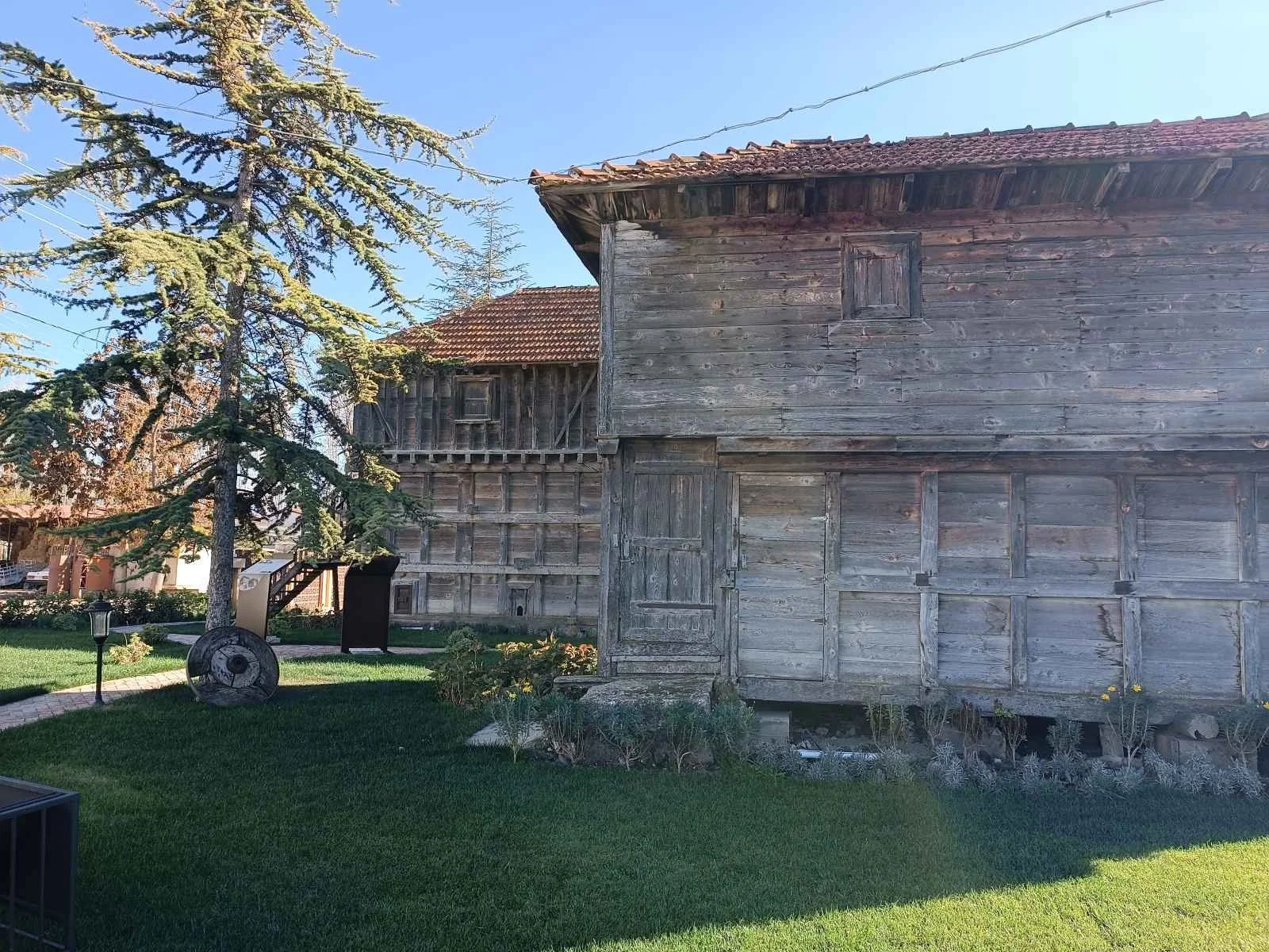 Ahşap eski bir ev ve önünde çimlerle çevrili bahçe bulunuyor, ağaç ve güneşli bir gökyüzü var.