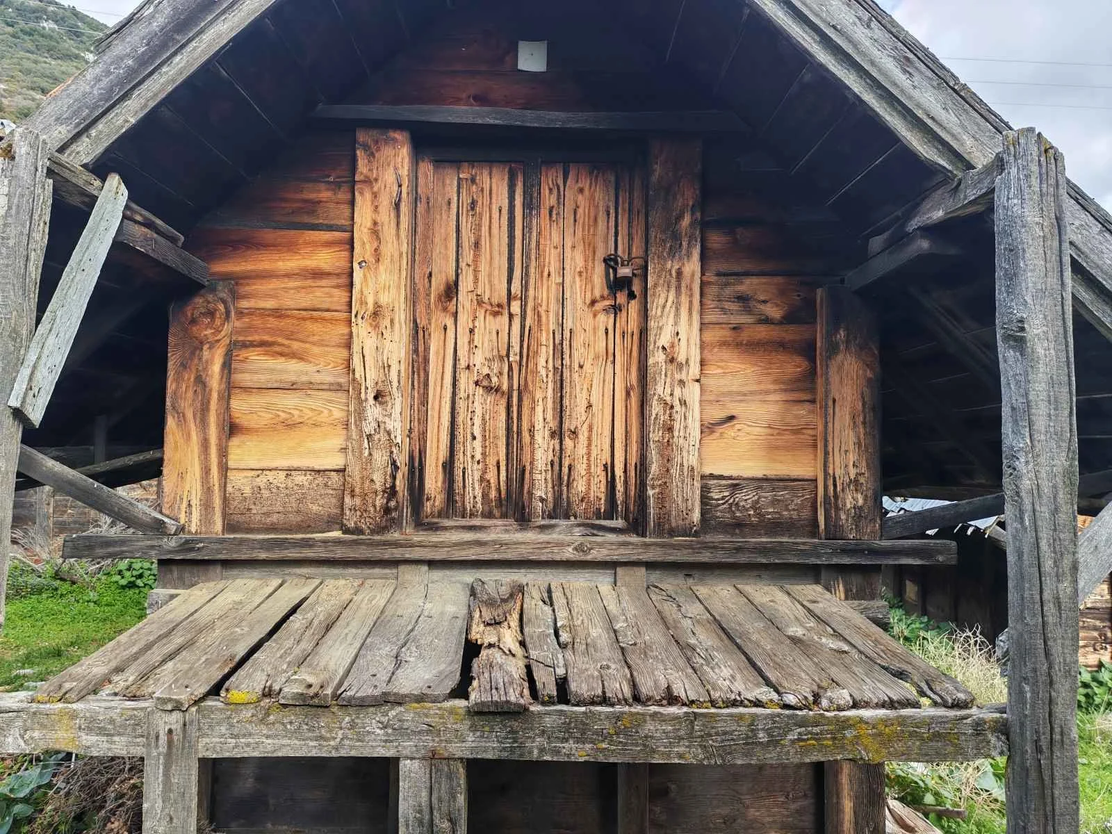 Ahşap eski bir kulübe görünümü, kapısı kilitli ve üzerinde çatlaklar var, etrafında yeşillikler ve dağlar bulunuyor.