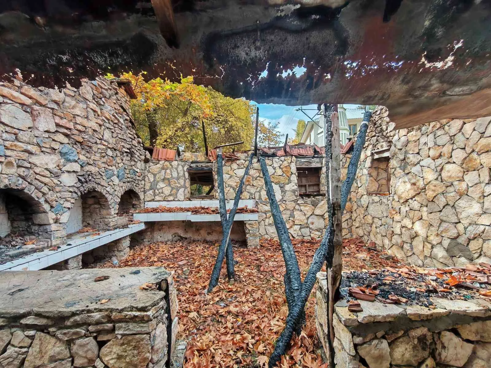 Yıkılmış taş duvarlar ve milletlerin ateş almış odunlar. Dış çekim, sonbahar yaprakları ve ağaçlar görünmekte.