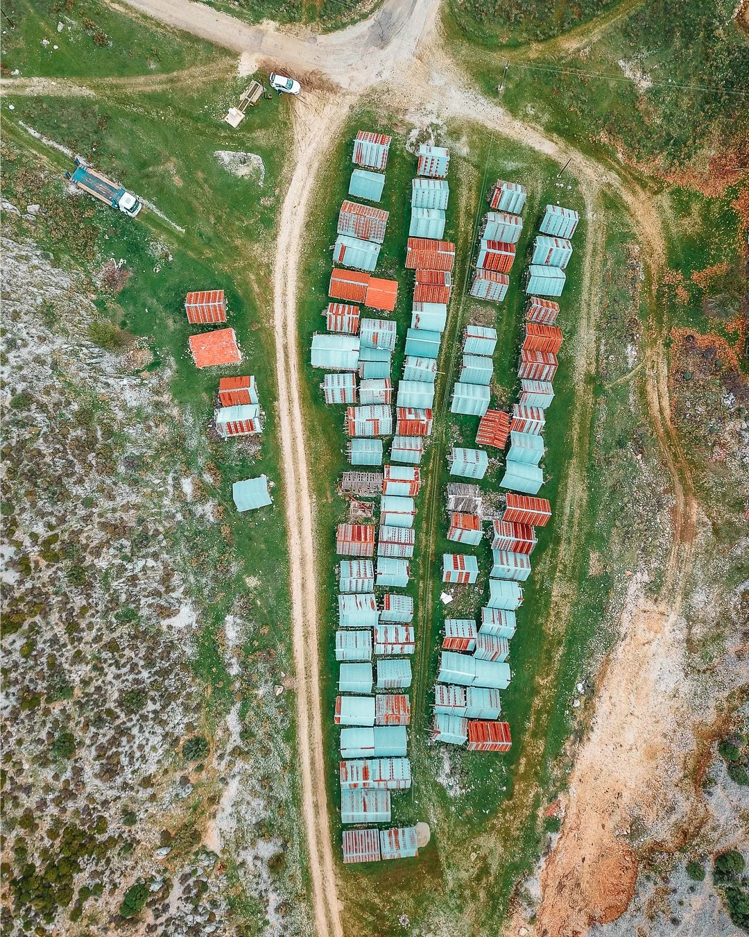 An aerial view of rows of small cabins with metal roofs, situated on a grassy and rocky landscape with dirt roads running through the area.