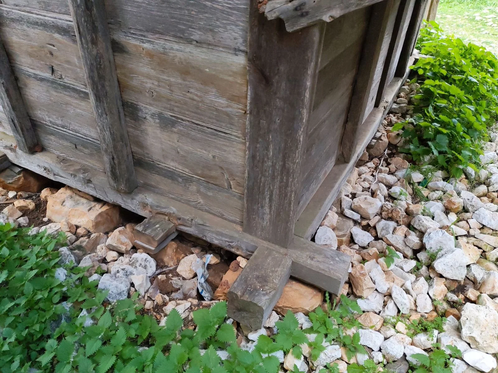 Ahşap bir yapının temel kısmı taşlar üzerinde duruyor, etrafında yeşil bitkiler bulunuyor.