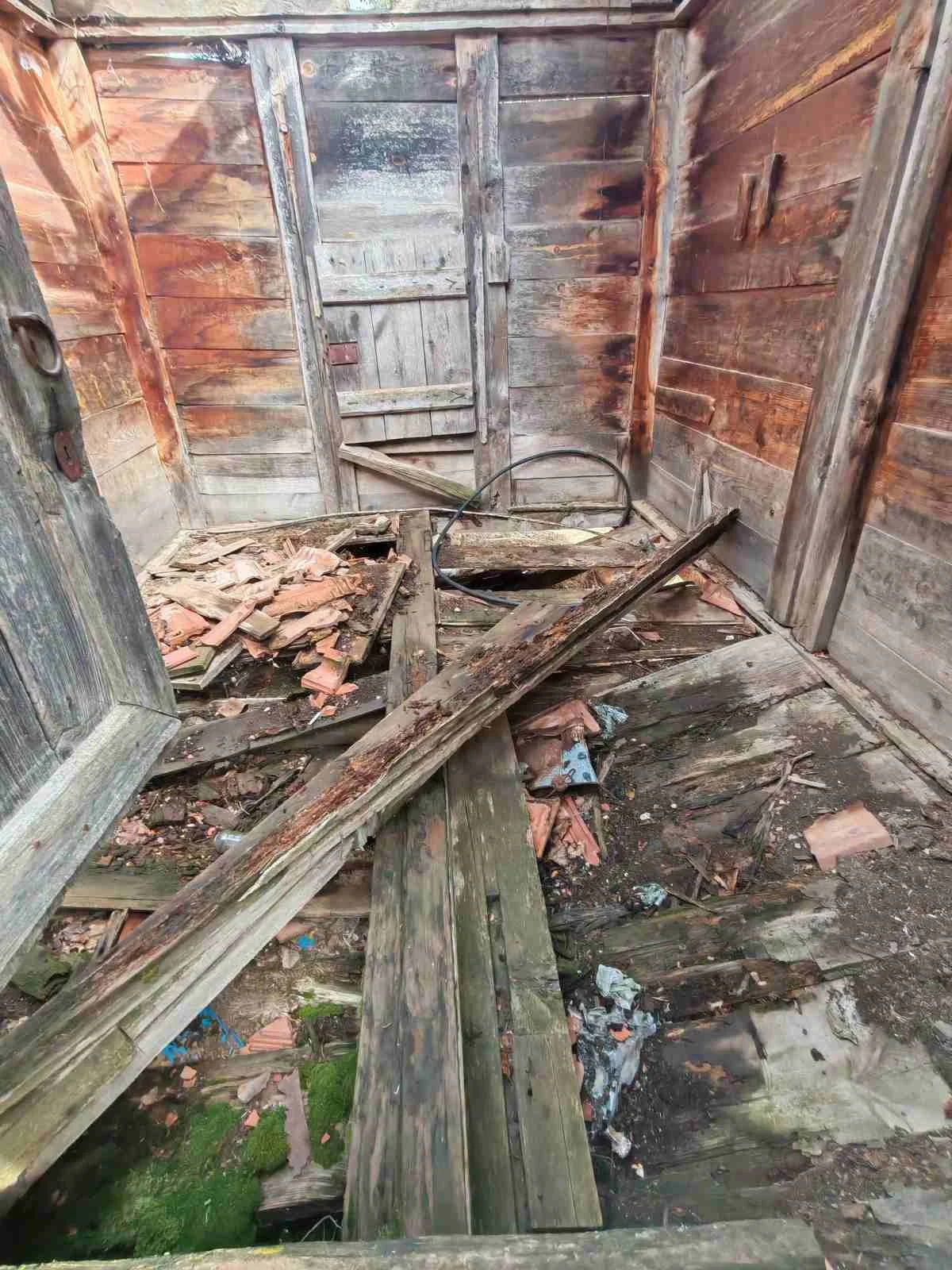 Yıkık ve dökülen ahşap bir oda, zemininde kırık çatıklar ve çökmüş yapı malzemeleri var.