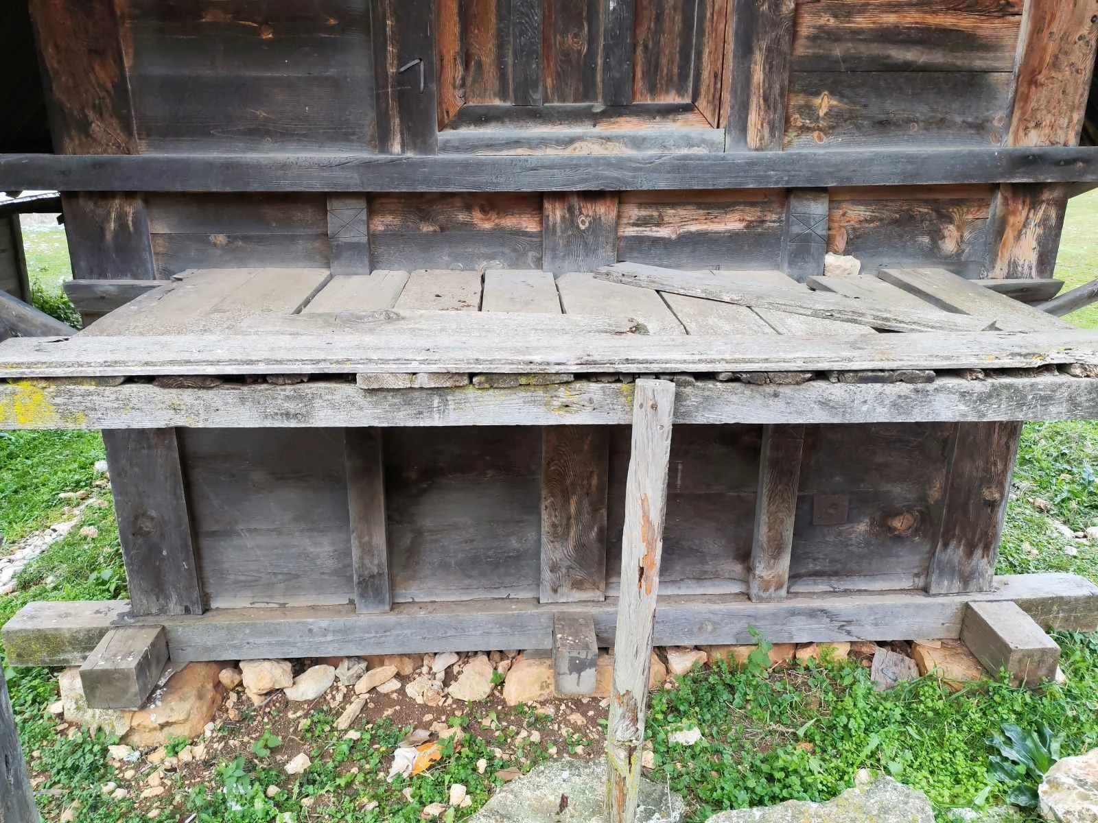 Eski ve yıpranmış ahşap bir yapı, altında toprak ve çakıllar, çevresinde yeşil çeşitli bitkiler var.