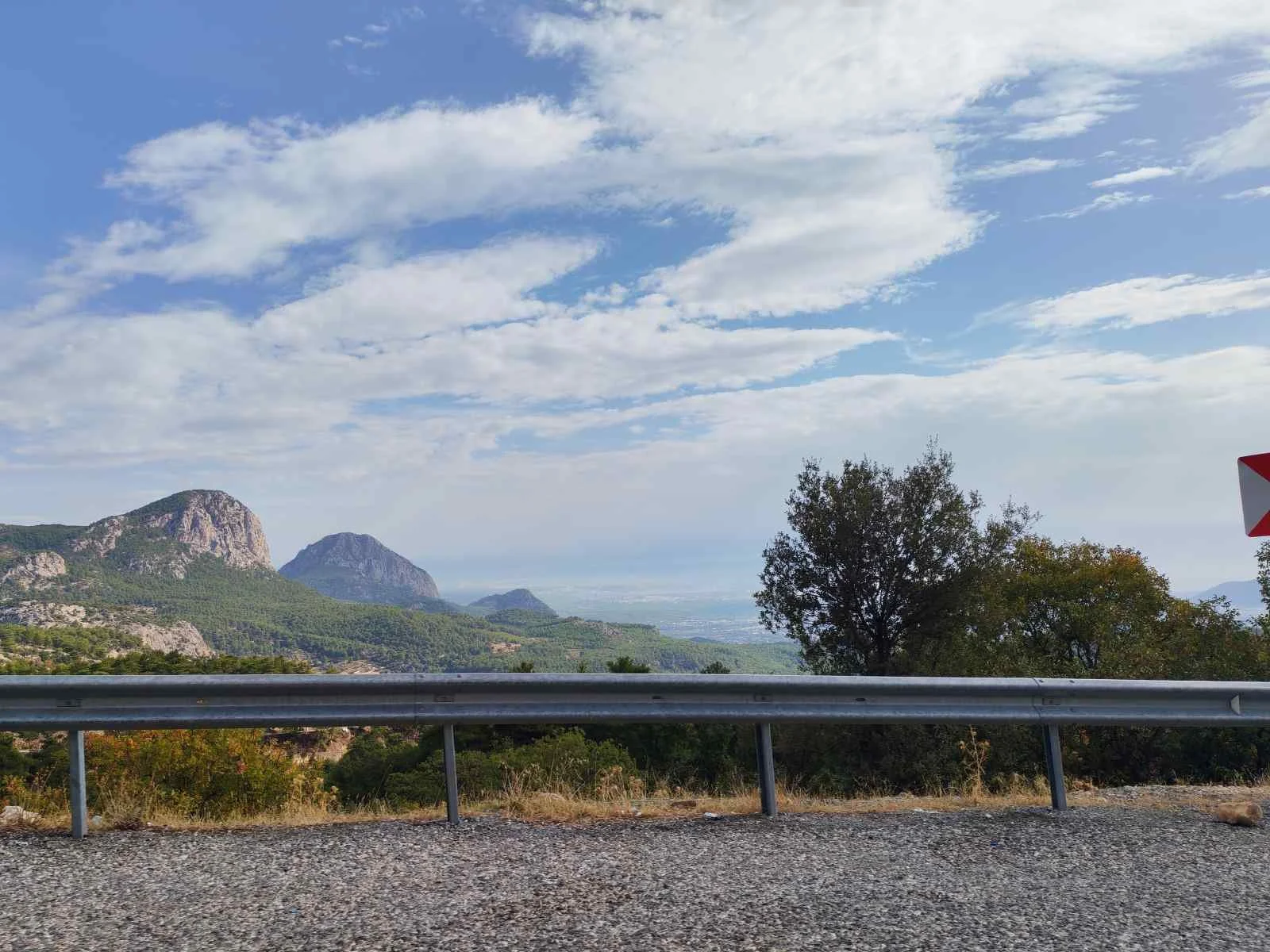 Dağlar ve ağaçlar ile mavi gökyüzü ve beyaz bulutlar, karayolu kenarında metal bariyer ve uyarı levhası ile açık manzara.