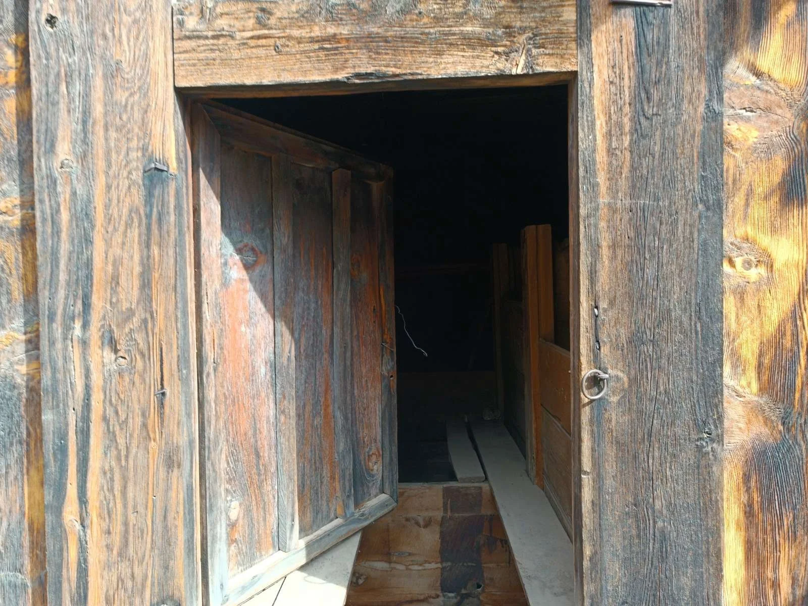 Ahşap bir kapı ve çerçevesi, açıkta duran iki kanatlı kapı, kapı eşiğinde tahta bloklar ve içeride karanlık bir alan.
