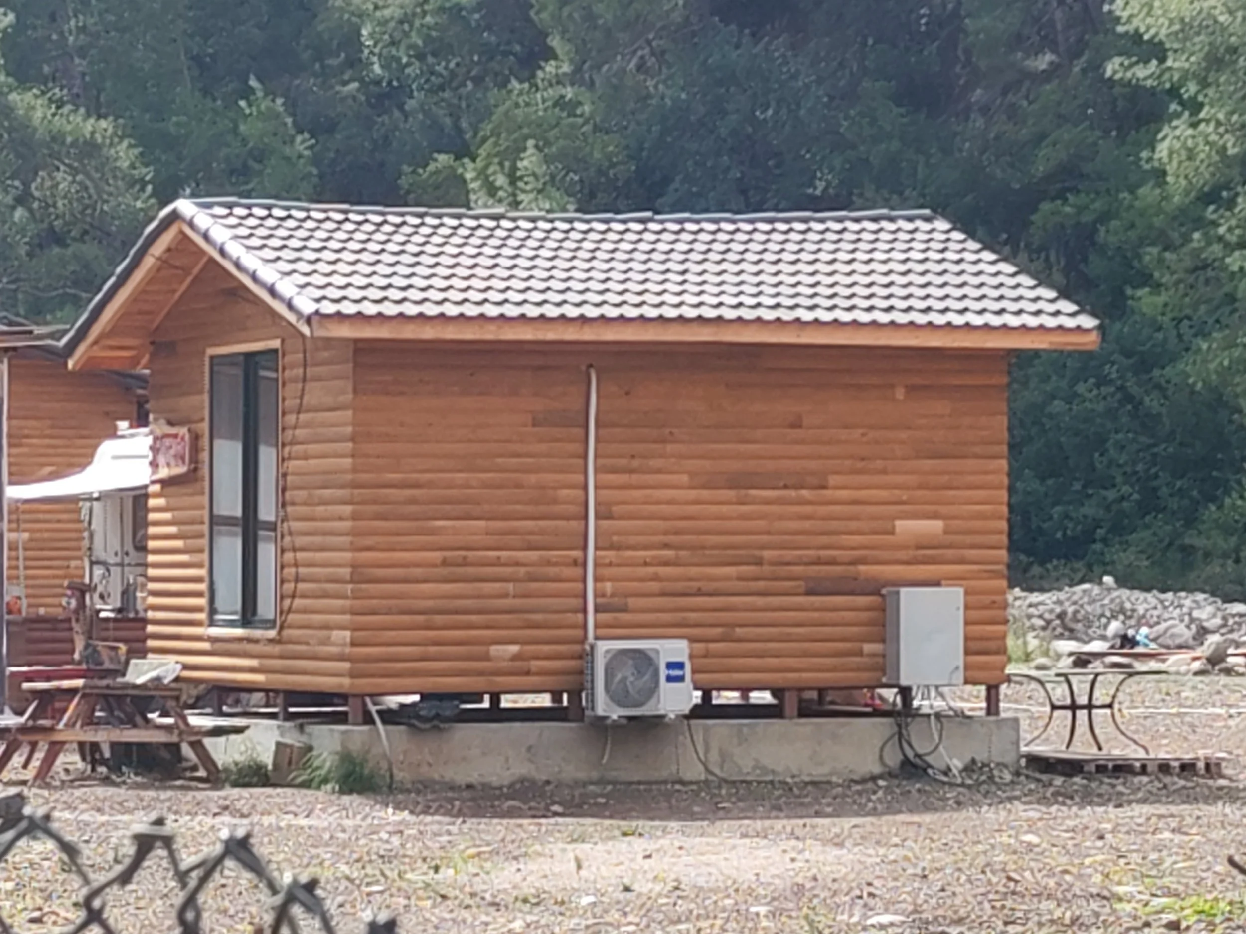 Ahşap ev, dış duvarlar log tarzında, küçük çatı ile kaplanmış, hava koşullarına dayanıklı malzeme ve elektrikli cihazlar dış ortamda yer alıyor, çevresi taş ve toprak ile çevrili, arka planda ağaçlar ve doğa bulunuyor.