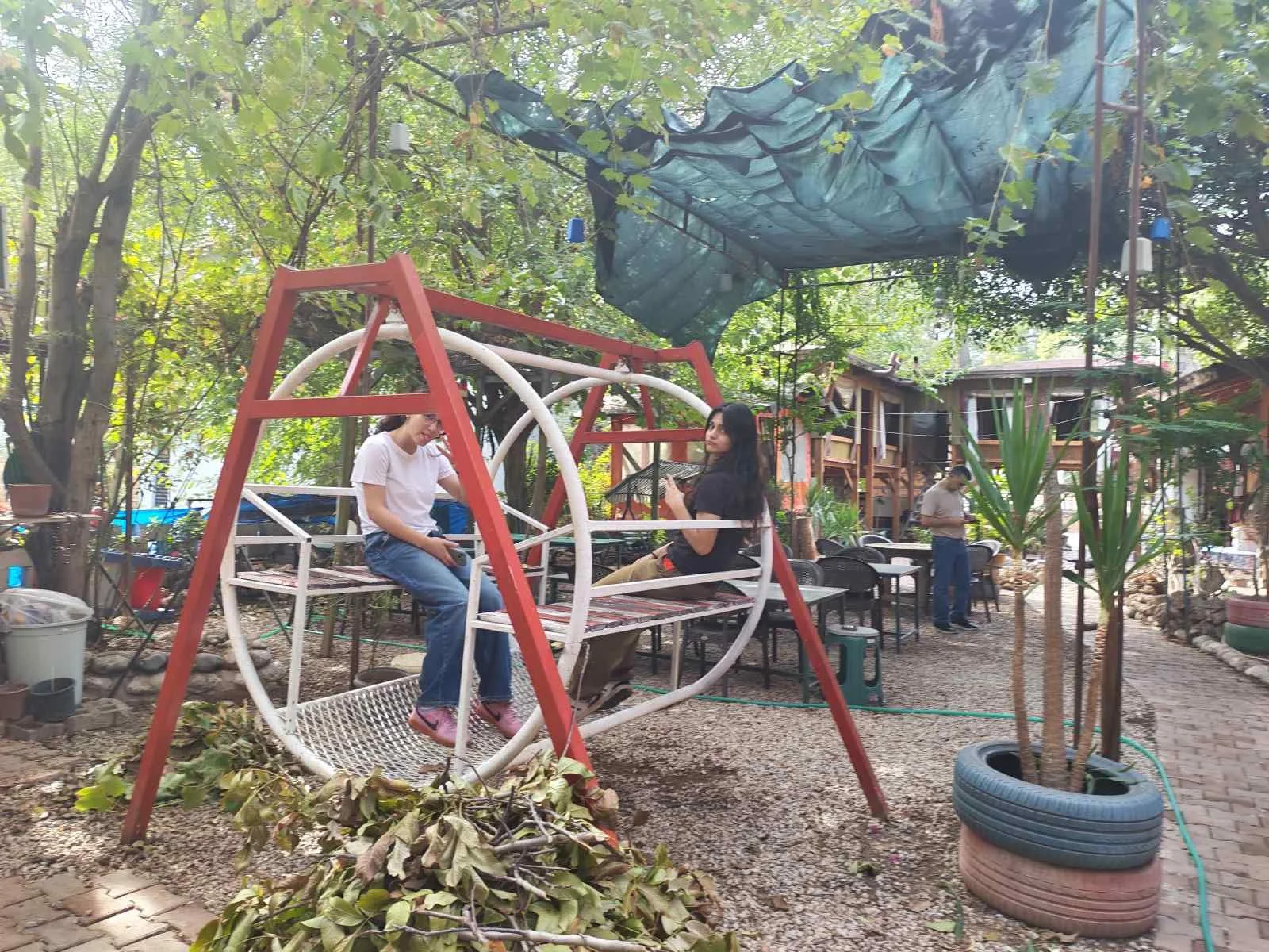 Bahçede çocuklar ve yetişkinler oyun oynuyor. Bir çocuk salıncağa otururken başka bir çocuk salıncağın çerçevesinde oturuyor. Arka planda birkaç yetişkin masa ve sandalye ile oturuyor. Bahçede ağaçlar ve bitkiler var, bazı bitkiler büyük saksılarda b