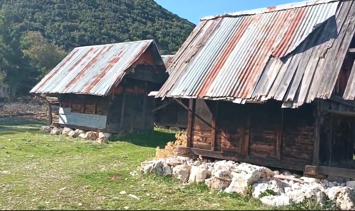 Ahşap ve galvaniz çatıya sahip eski kulübeler veya evler, taş temeller üzerinde, yeşil alan ve ağaçlar ile doğa manzarası.