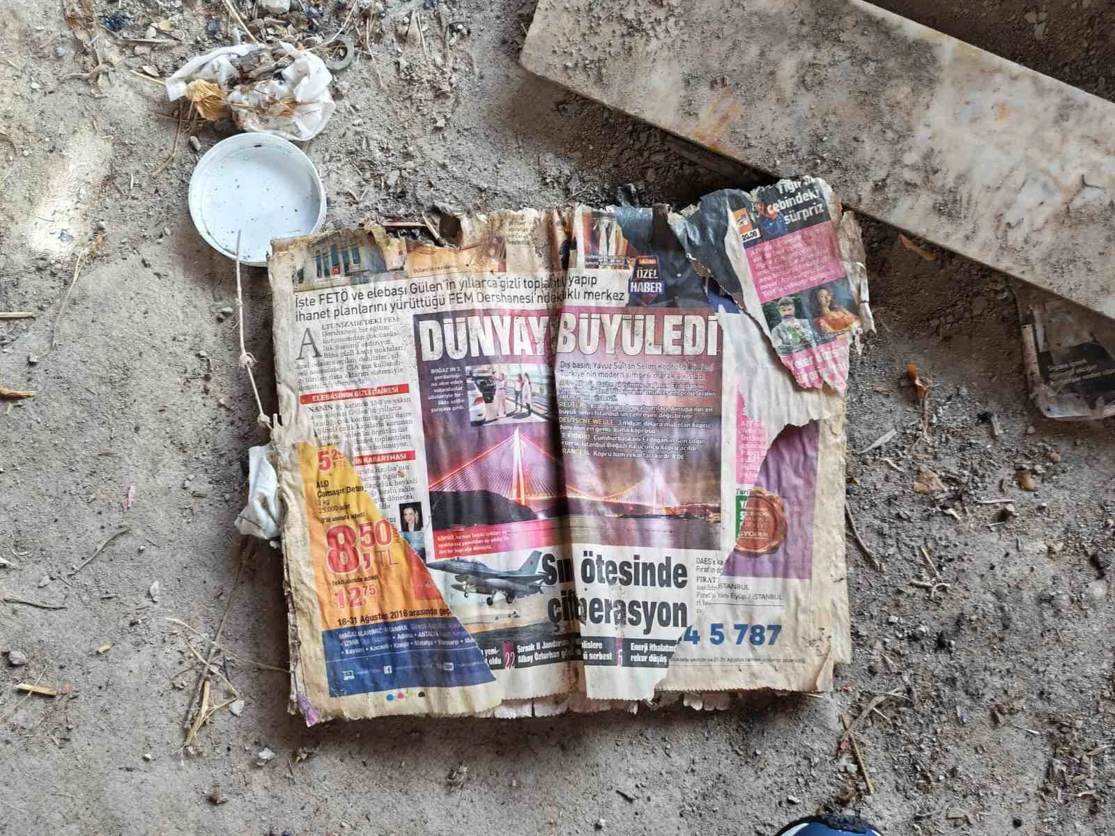 Tozlu zemin üzerinde eski, yırtık ve katlanmış bir Türkçe gazetesi, yanında bazı metal ve plastik nesneler ve bir içecek kalıntısı ile birlikte. Gazetenin biraz üst kısmı taş veya beton parçası üzerine yerleştirilmiş.