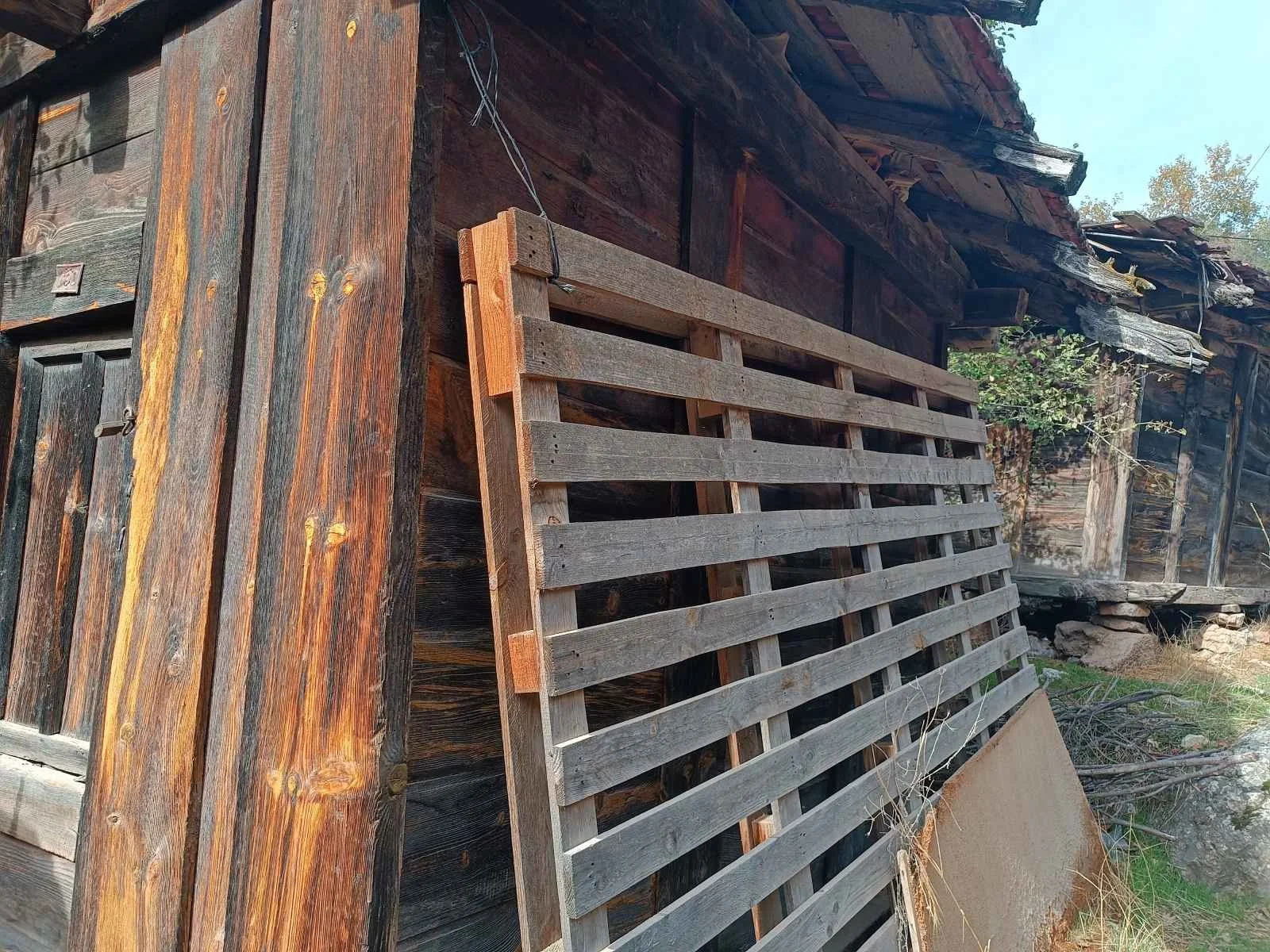 Eski ve ahşap bir evin dış cephesi, yan duvarında yatay ve dikey ahşap kirişler ve ızgara şeklinde bir ahşap parmaklık yer alıyor, arka planda ağaçlar ve gökyüzü görünmekte.