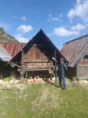 İki kişi, geleneksel ahşap kulübe önünde duruyor, açık gökyüzü ve dağlar arka planda.