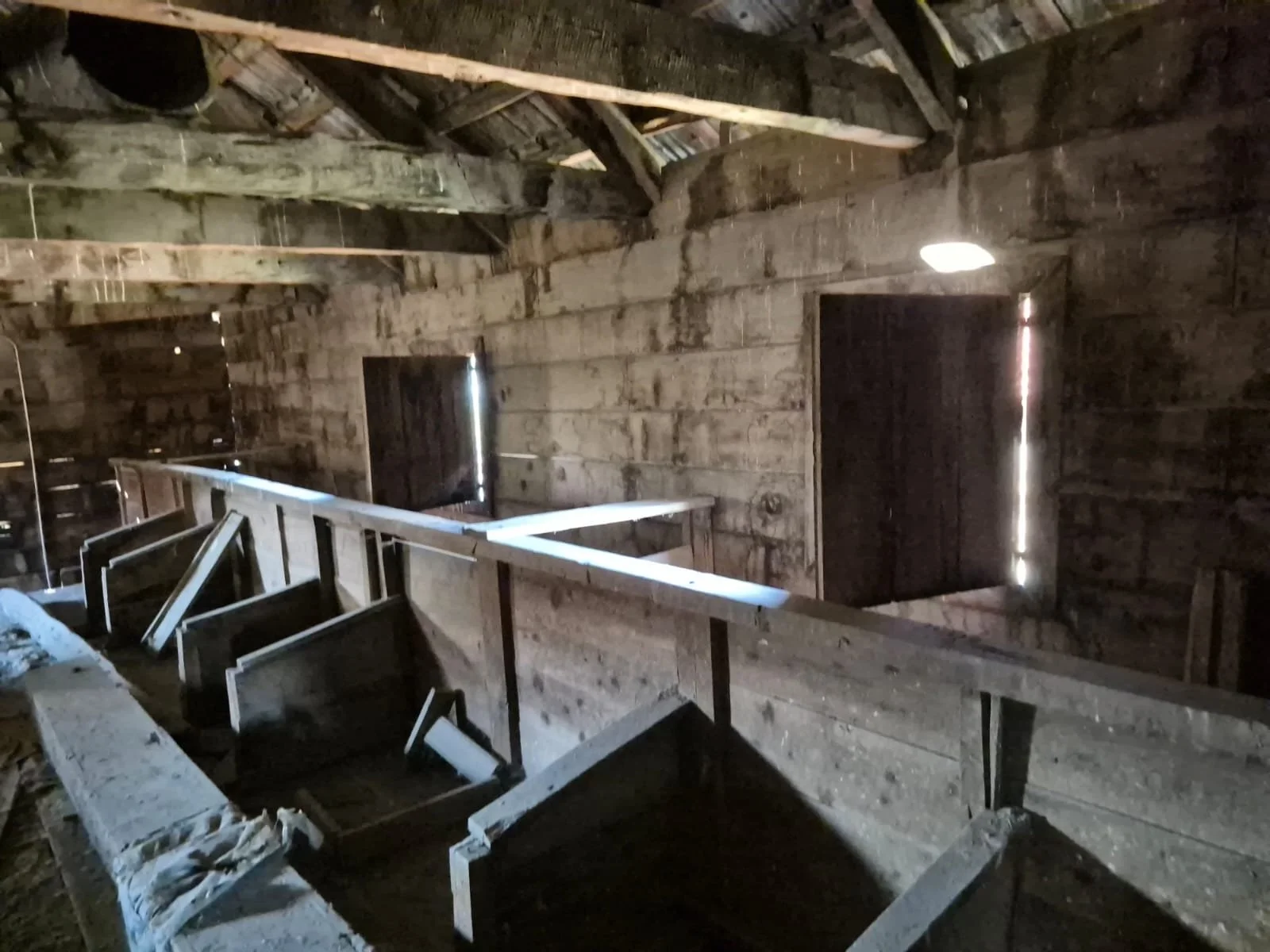 Ahşap bir inşaat veya restore edilmekte olan eski bir bina içi, duvarlar ve tavan ahşaptan ve doğal ışık küçük açıklıklardan içeriye sızıyor.