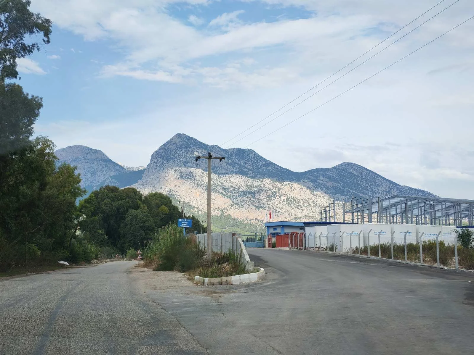 Dağlar ve gökyüzü arka planda, asfalt yol ve bazı binalar ve elektrik direkleri ön planda görülen bir kırsal alan.