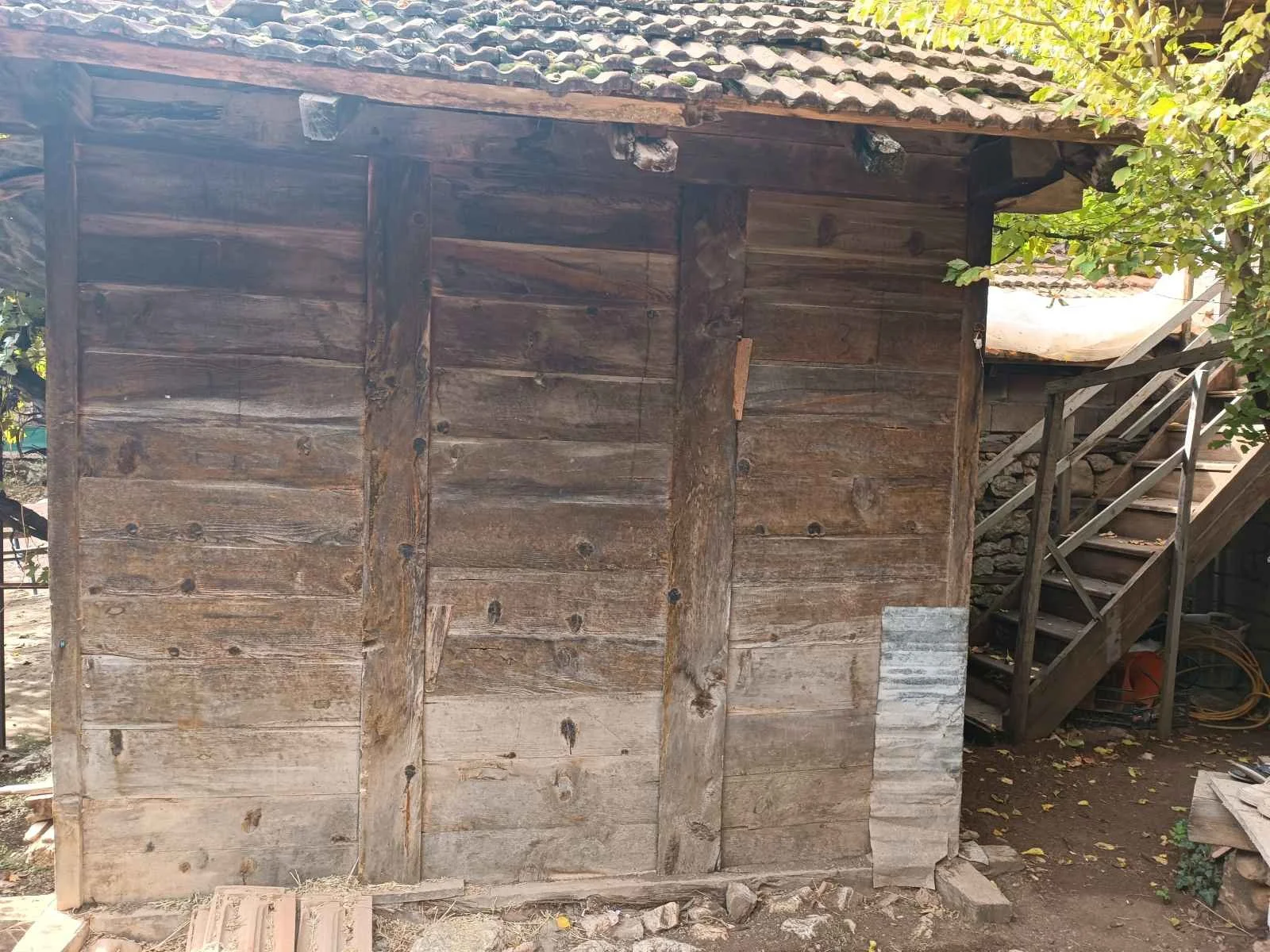 Ahşap bir kulübe veya yapı, dış duvarları gösteriyor, kapı veya pencere yok, dışısı doğal ahşap ve taş bariyerlerle çevrili, arka planda ağaçlar ve merdiven var.