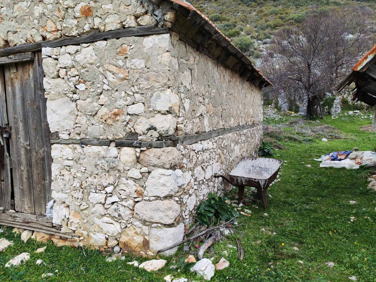 Kırsal alanda taş duvarlı küçük yapı ve çevresinde bir el arabası, yeşil alan, ağaçlar ve dağlar görünmektedir.