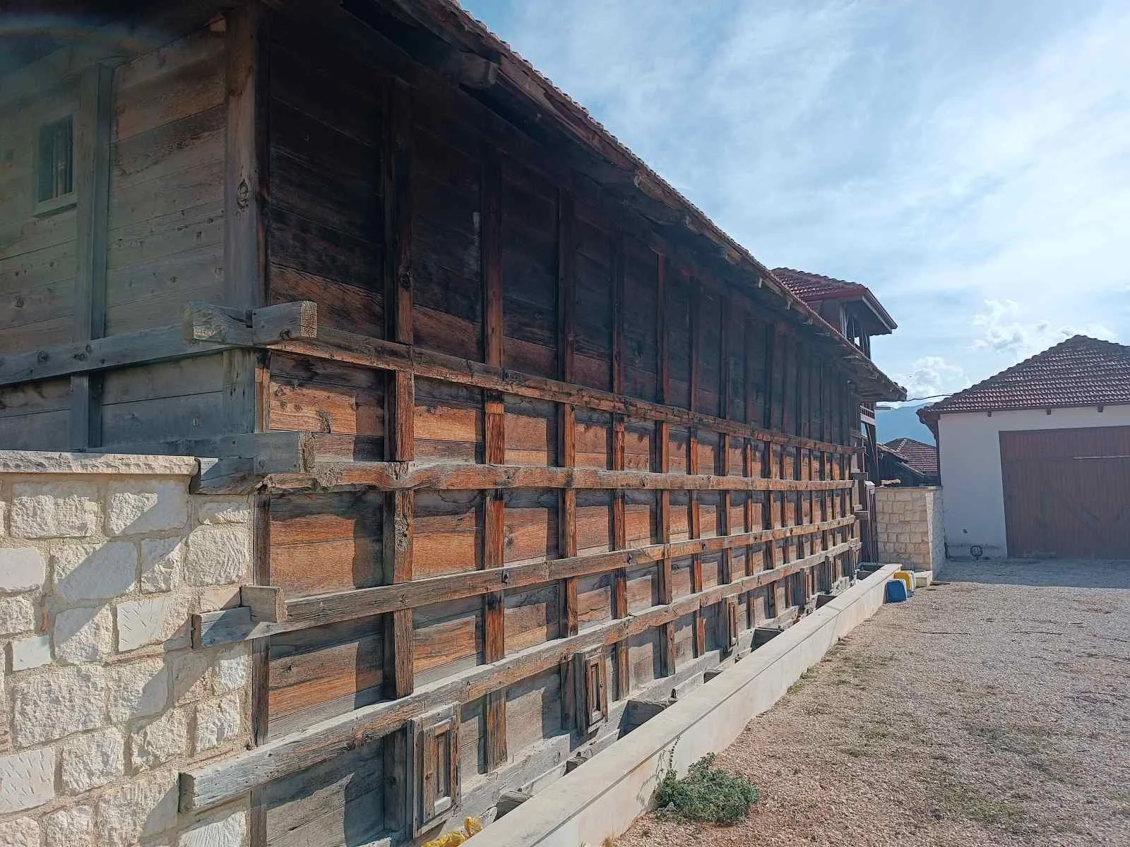 Bir inşaat alanında, ahşap çerçevesi bulunan ve beton temel üzerinde yer alan eski ahşap yapı, arka planda mavi gökyüzü ve birkaç modern yapı görünmekte.