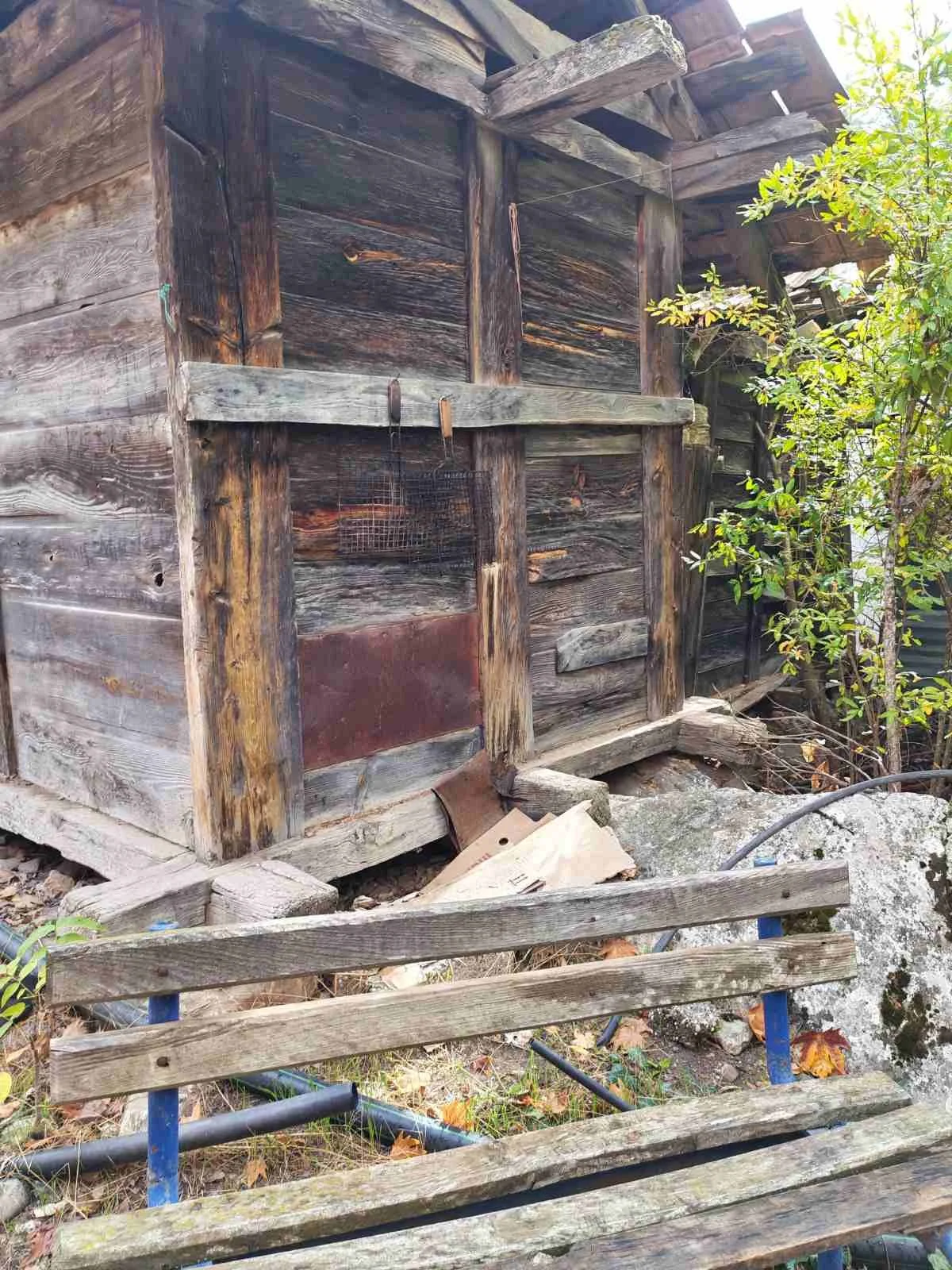 Eski ve bakımsız ahşap kulübe. Kulübenin önünde yıkılmış park bankı ve çeşitli atıklar var. Kulübenin yanında küçük bir ağaç bulunuyor.