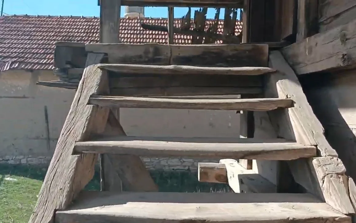 Ahşap merdiven, bir bahçede veya dış mekanda yer alıyor, arka planda kırmızı kiremitli çatılar ve duvarlar görülüyor.