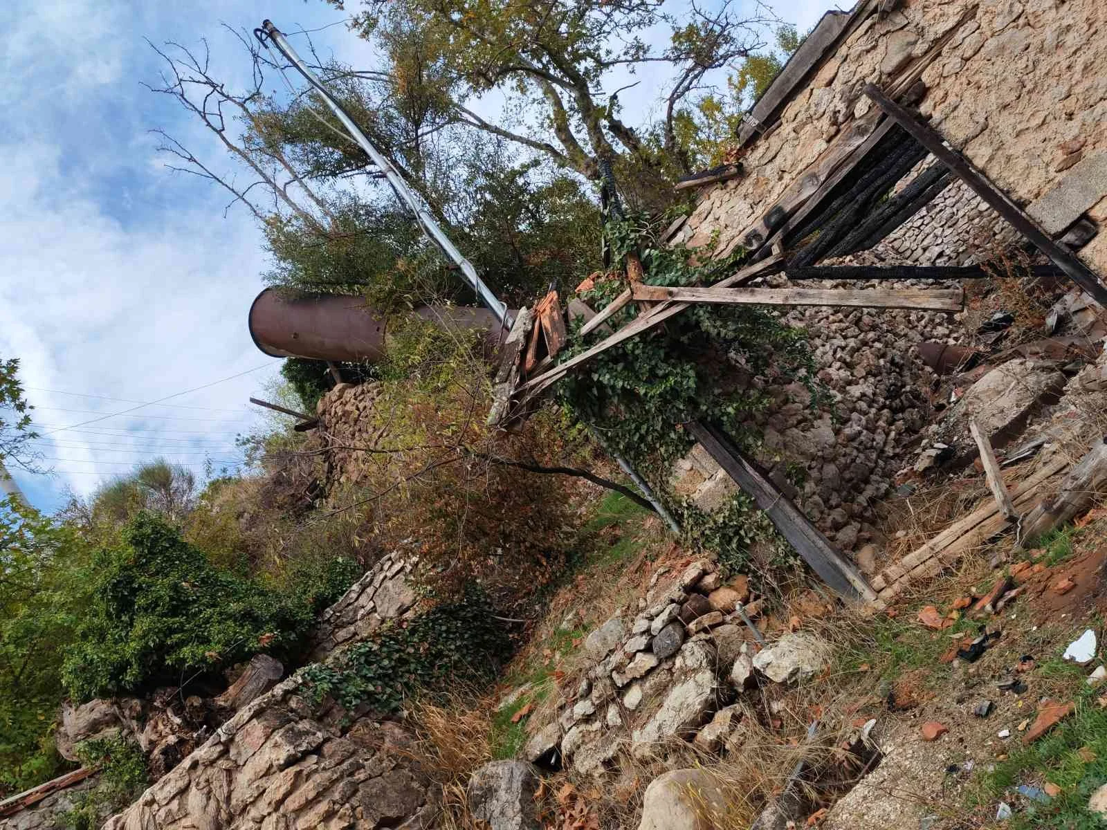 Açık mavi gökyüzüne karşı, yıkık bir duvar ve eğilmiş bir yapı ile etrafa yayılmış dallar ve yapraksız ağaçlar, terkedilmiş veya hasar görmüş bir alan görünümü.
