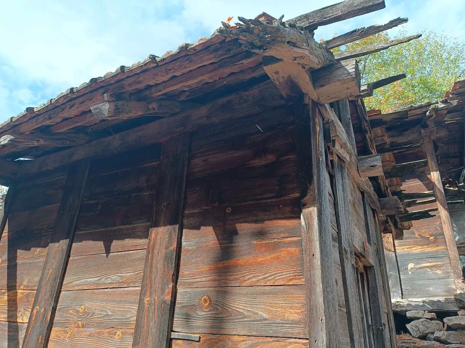 Ahşap eski bir yapı, çatısı ve duvarları ahşap plakalardan yapılmış, bir kısmı bozulmuş ve çatlamış.