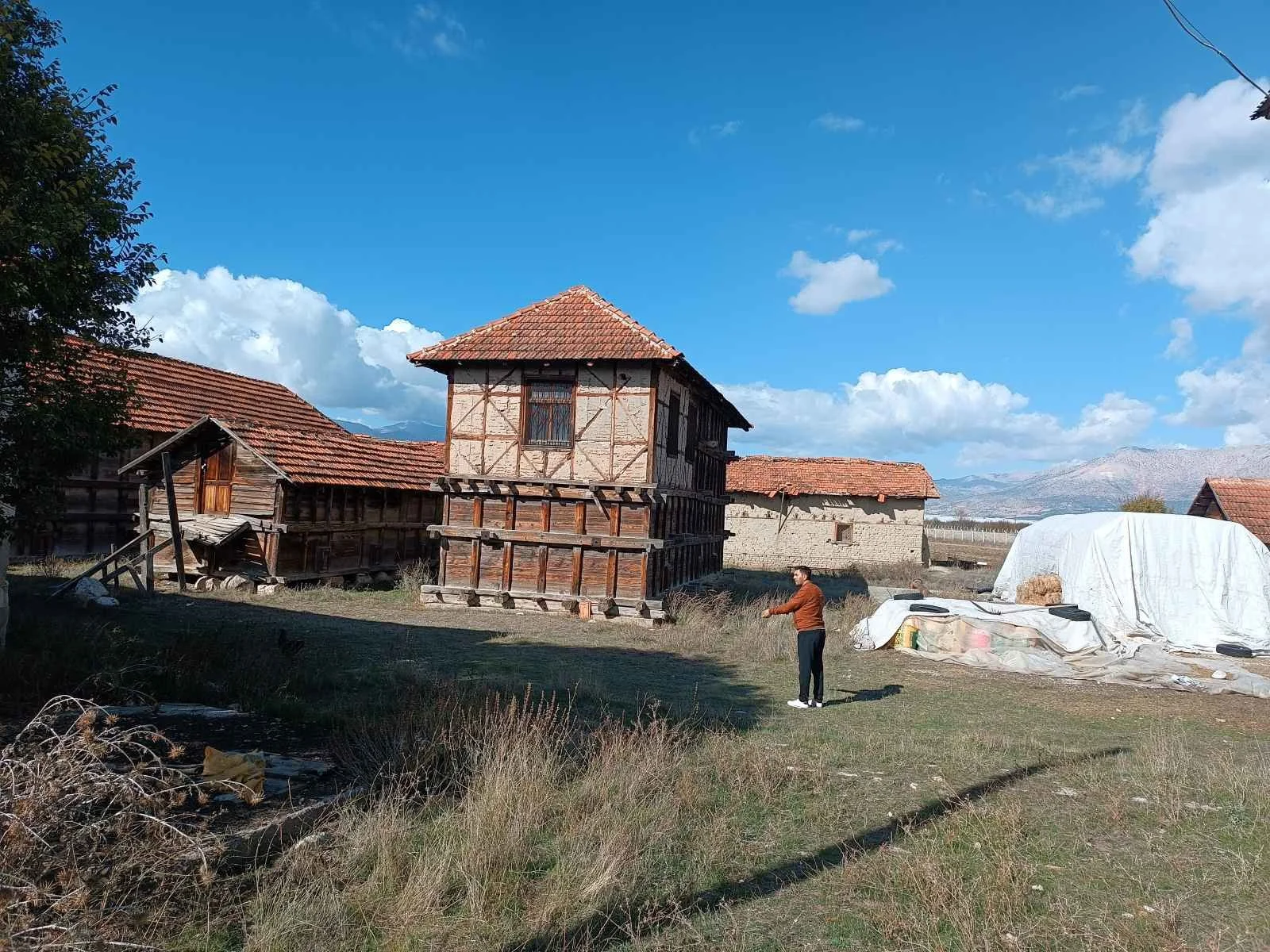 Kırsal alanda, birkaç eski taş ve ahşap yapıya sahip evler, açık gökyüzü ve dağlar, bir kişi ayakta dururken ve etrafta çeşitli yapılar ve doğal çevreler görünmektedir.