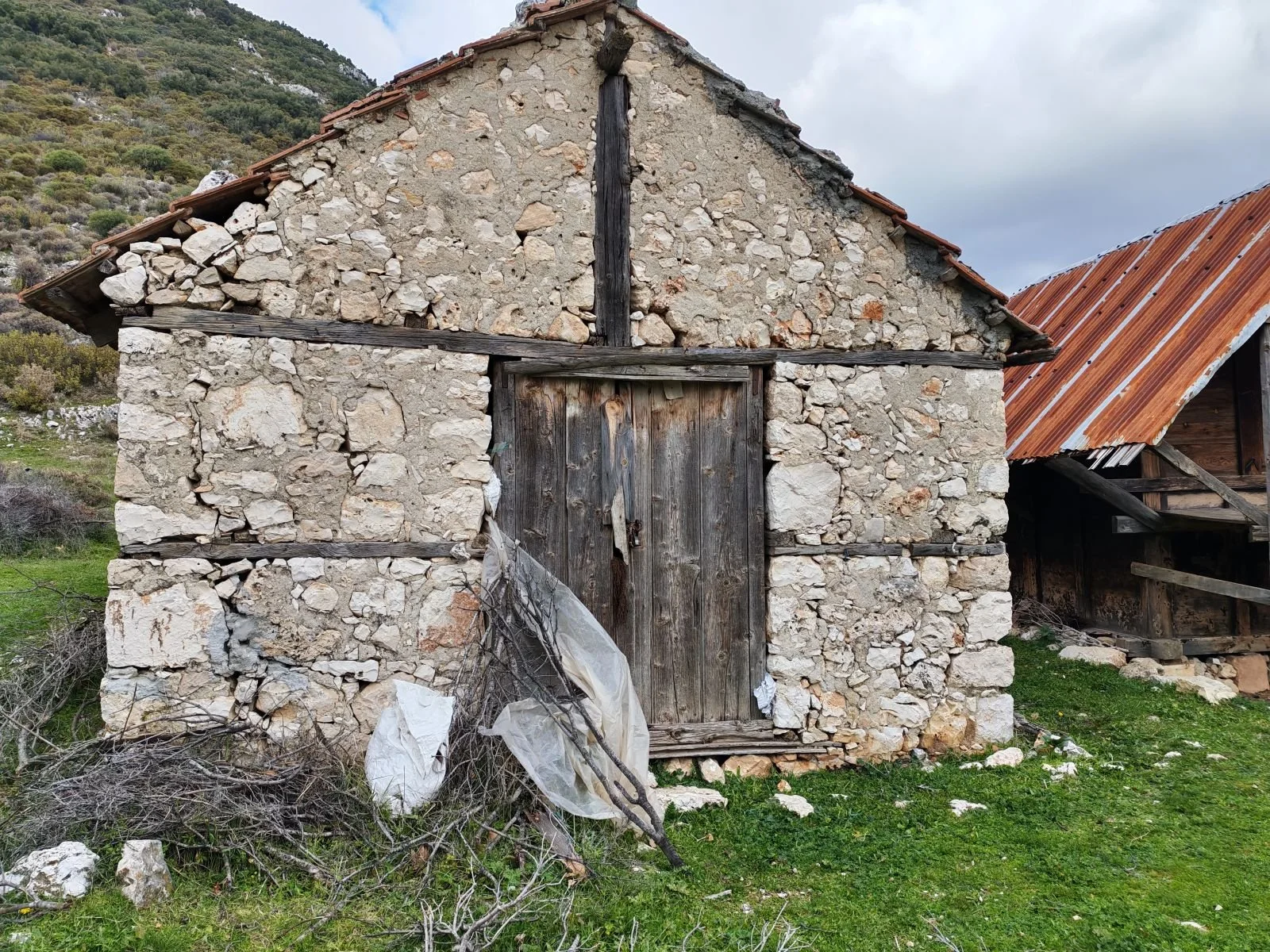 Taş ve ahşaptan yapılmış eski bir kulübe, önünde çalılık ve toprak alan. Arka planda dağlar ve gökyüzü var.