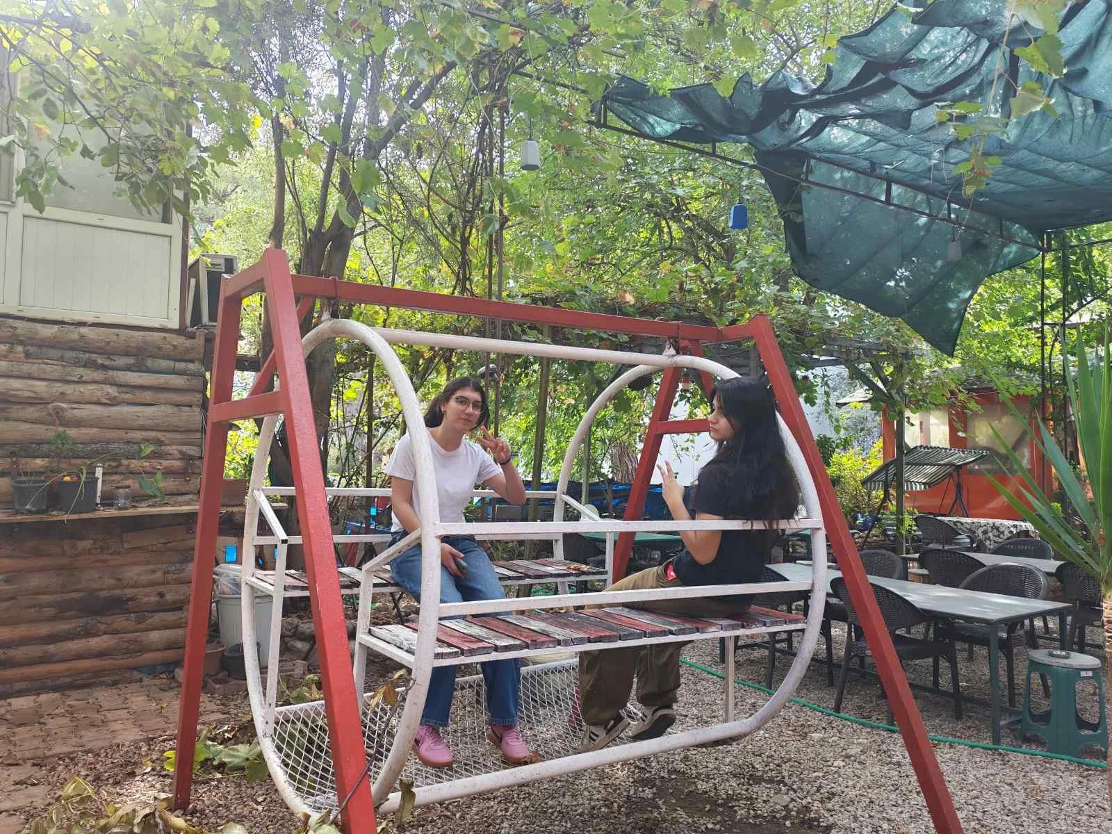 İki kadın, salıncağa oturmuş, doğa içindeki açık alanda sohbet ediyor.