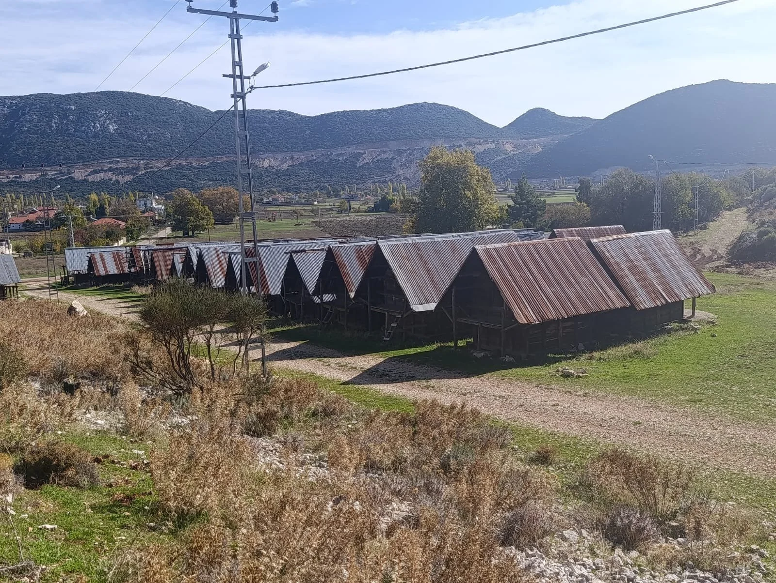 Dağlarla çevrili kırsal alanda, metal çatıya sahip ahşap yapılar sıralanmış.