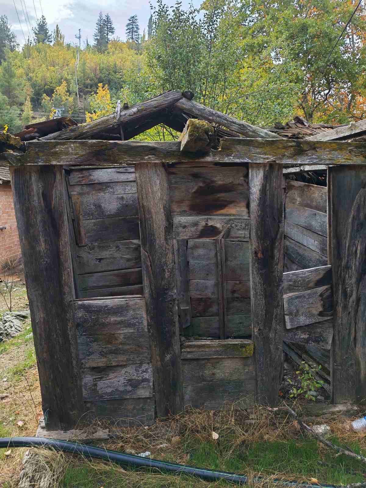 Ahşap malzemeden yapılmış, yıkılmış bir bina. Çatısı ve duvarları hasar görmüş ve eski. Çevresinde ağaçlar ve doğal manzara var.