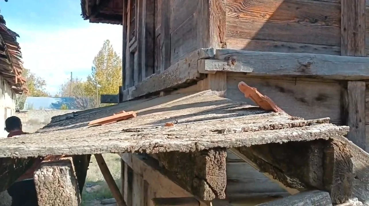 Ahşap yapılı eski bir ev veya kulübe, çatısı ve yan duvarları görülüyor, bazı kırık ve kullanılmayan çatı kiremitleri yerde duruyor.