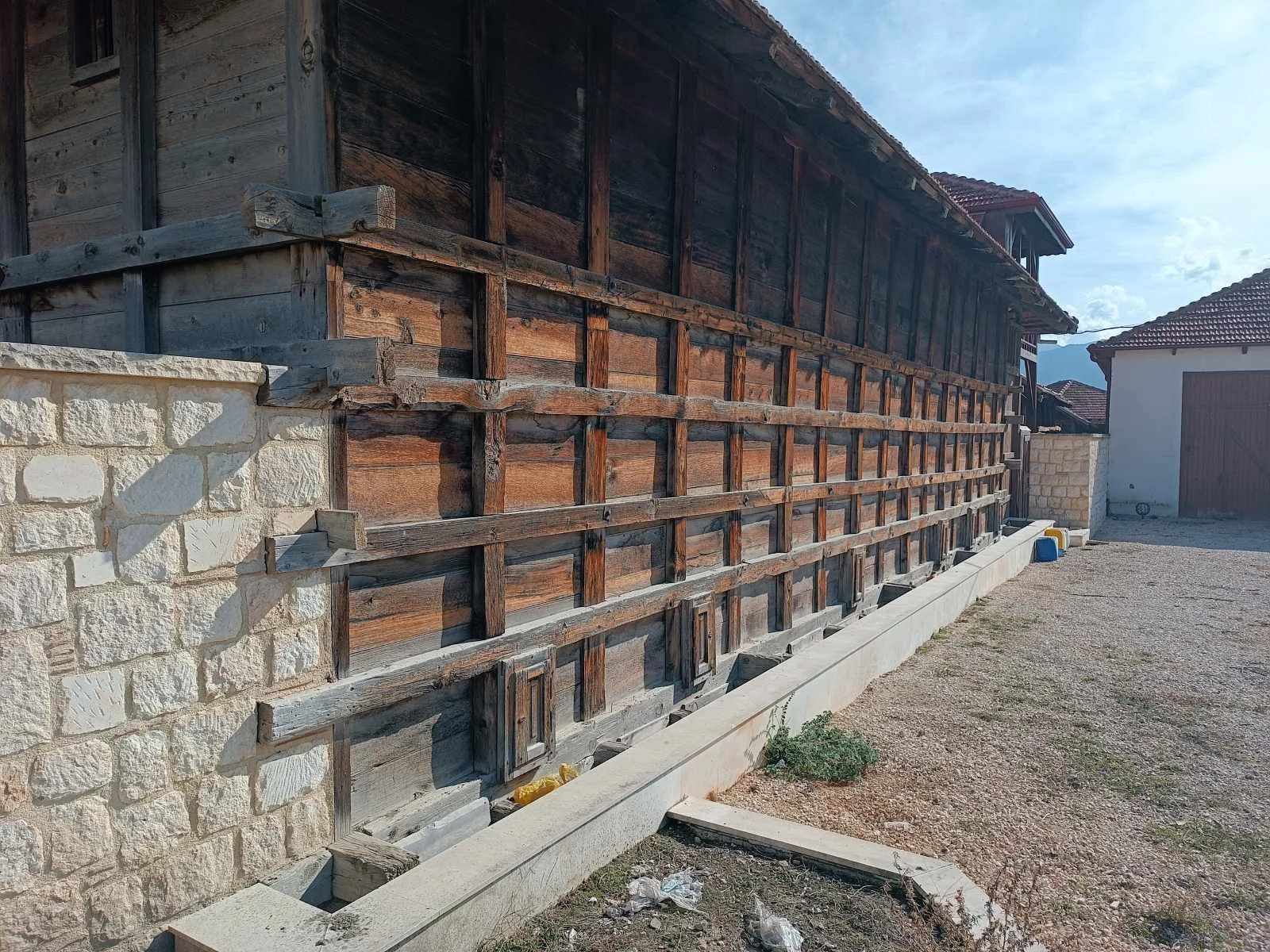 Ahşap çatılı ve duvarlı bir bina önü. Çevre toprak ve taş dolu, yanlarda birkaç küçük araç yer alıyor.