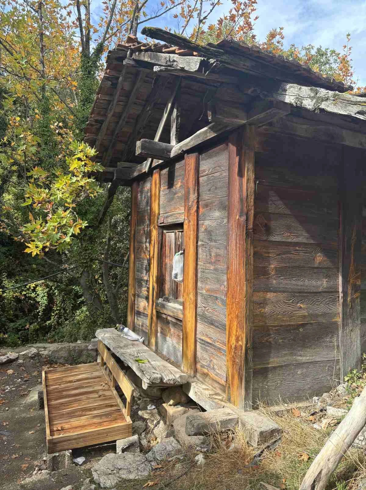 Ahşap bitkiyle çevrili, eski ve küçük bir kulübe veya kulube. Çatı yapısı ahşap ve kırık. Kulübenin önünde taştan ve ahşaptan yapılmış küçük bir oturma alanı ve yanına yerleştirilmiş birkaç plastik şişe var. Çevrede ağaçlar ve kuru yapraklar bulunuyo