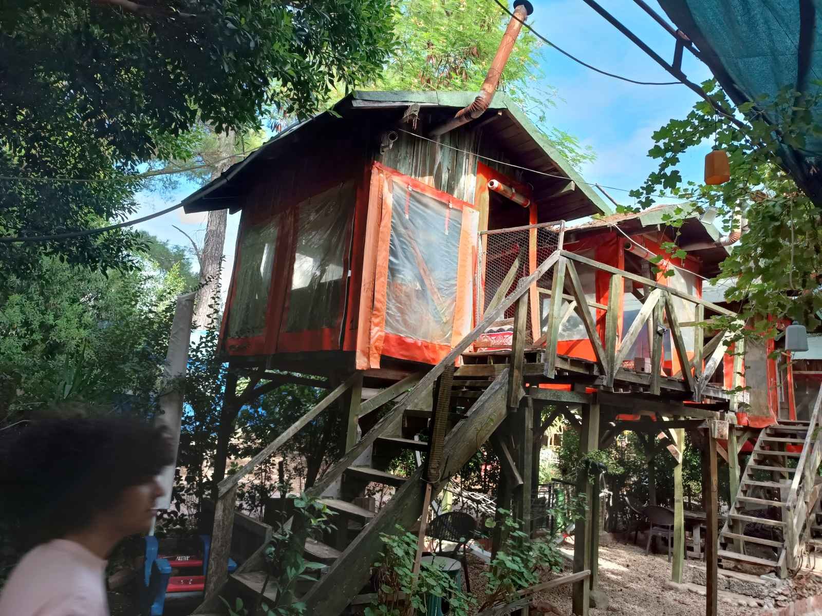 Ağaçların arasında yükseltilmiş ahşap bir ev, çatı ve duvarlar kısmen kaplanmış plastik malzeme ile. Çocuklar ve bahçe mobilyaları görünmekte.
