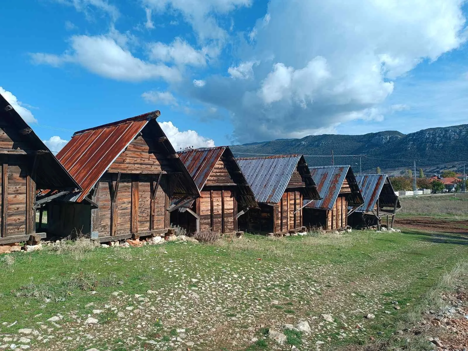 Eski, ahşap ve metal çatılı kulübeler, açık alanda, dağ manzaralı, gökyüzü mavi ve bulutlu.