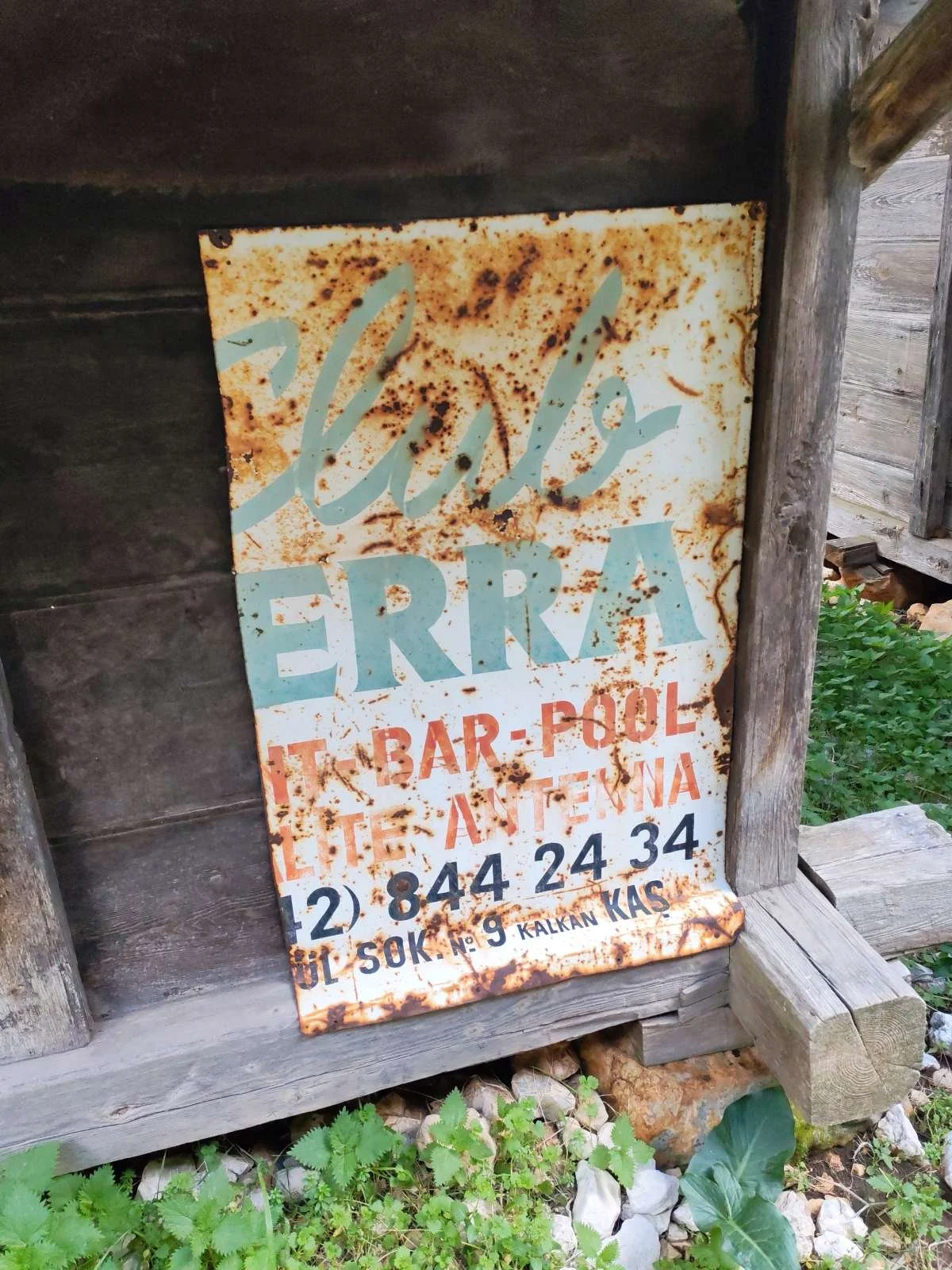 Paslı, eski metal levha, üzerinde mavi ve kırmızı harflerle yazılmış, ilk bakışta kafe veya bar bilgisi içeren ve çevresinde ahşap yapılar ile yeşillik bulunan dış mekan detayları.