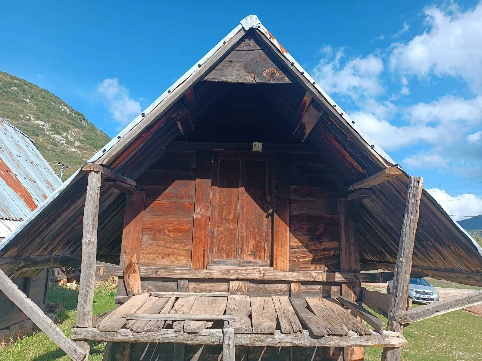 Odun kaplamalı ve ahşap çatıya sahip küçük ahşap kulübe veya ev. Önde bir verandası var, arka planda dağlar ve gökyüzü görülüyor.
