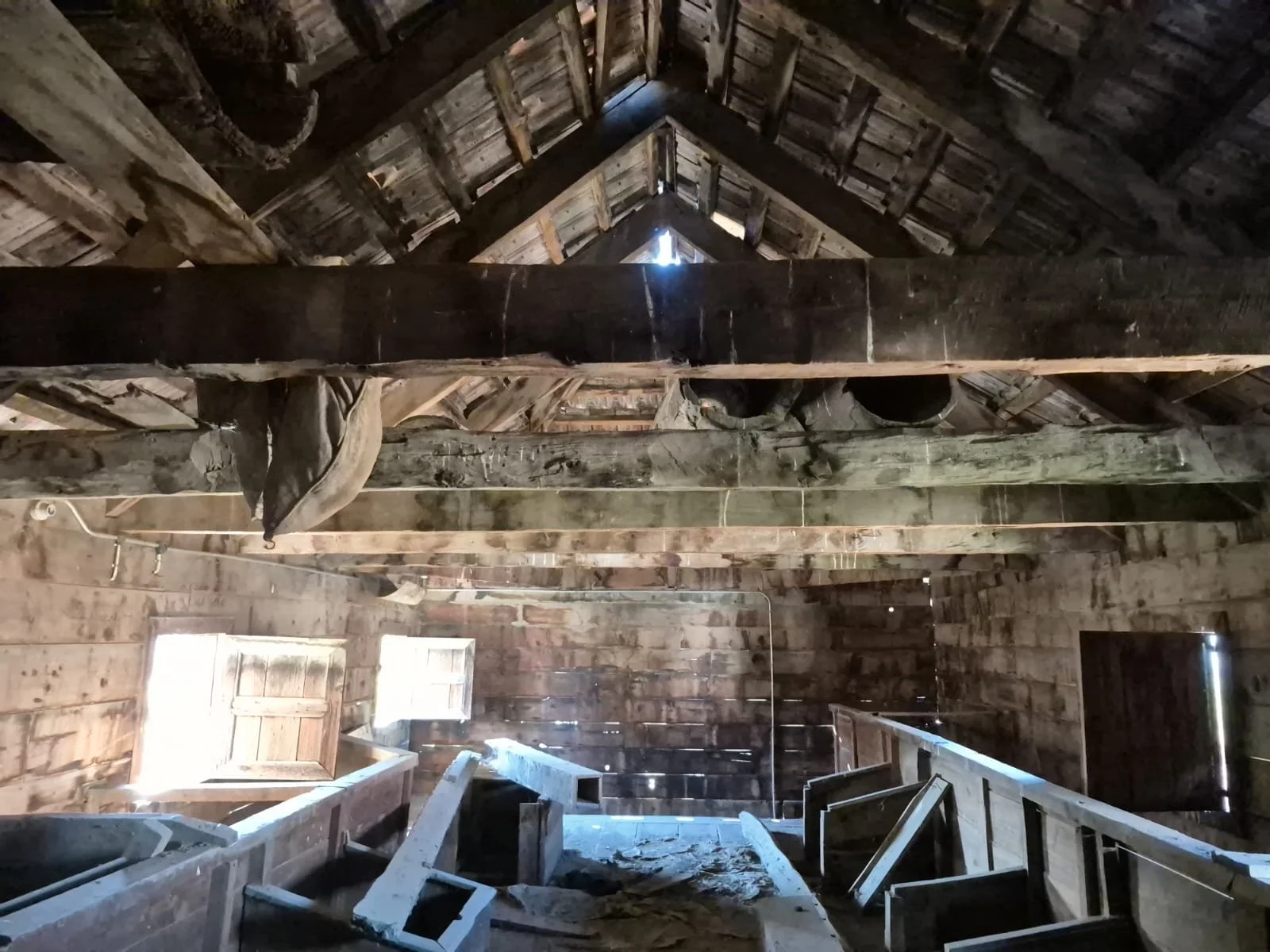 Eski ve ahşap bir yapının iç mekanı, tavan ve duvarlar odunlardan yapılmış, çatıda boşluk ve gün ışığı görülen, bazı eski eşyalar ve tahta merdivenler içeriyor.