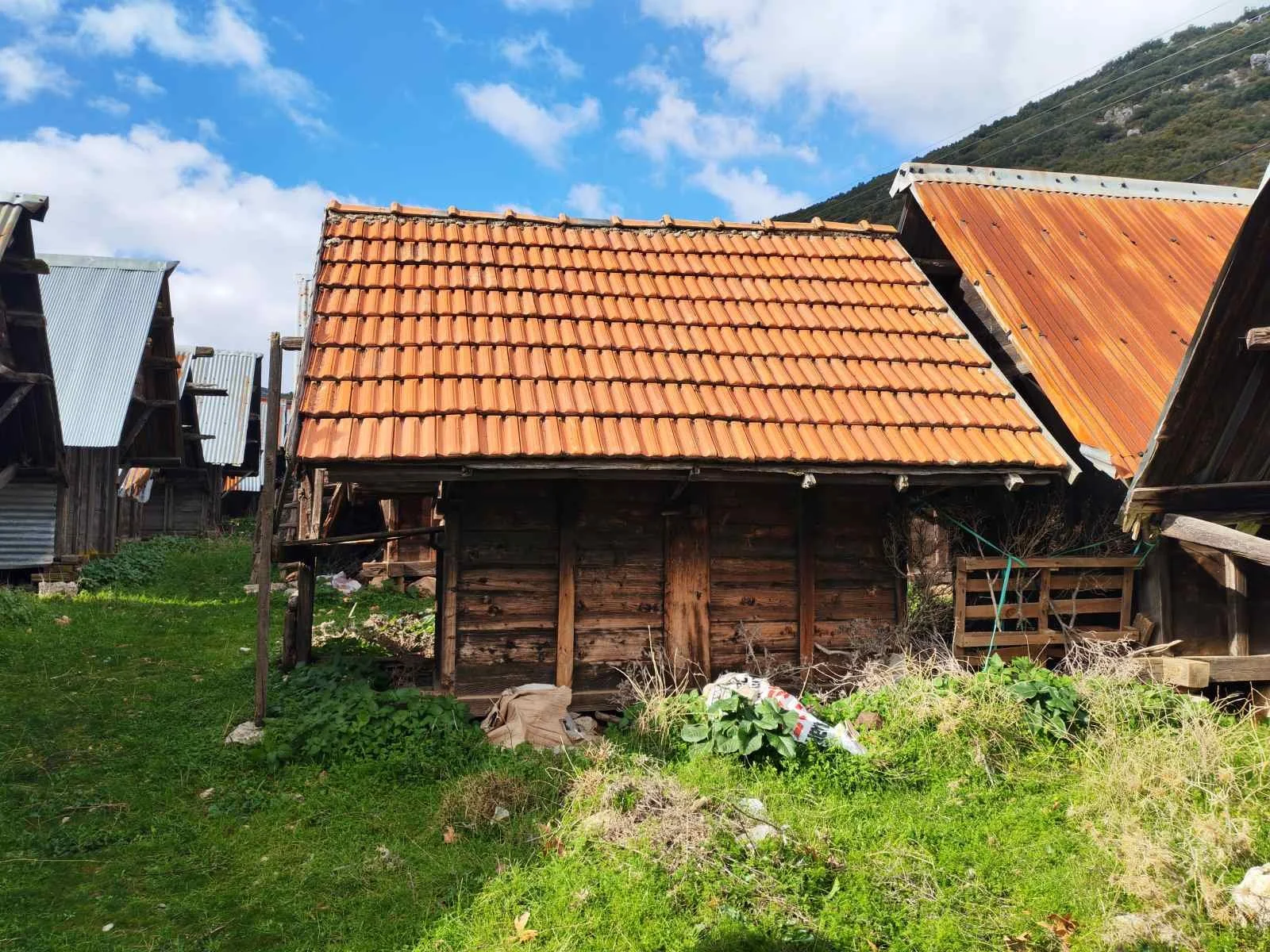 Eski ahşap ve kiremit çatılı küçük bir kulübe, etrafında yeşil çim ve diğer ahşap yapılar var, arka planda dağlar ve gökyüzü görülüyor.