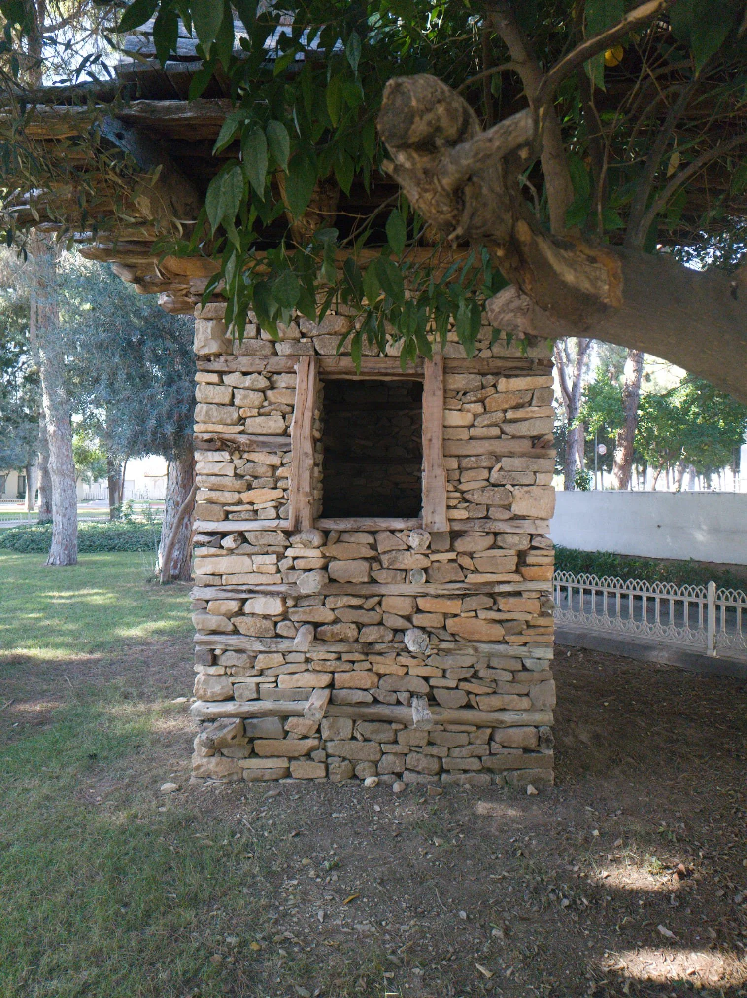 Küçük taşlardan inşa edilmiş, boş bir pencereye sahip taş kulübe, ağaçların gölgesinde, parkta yer alıyor.