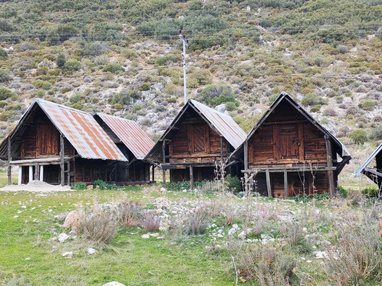 dörd tane ahşap, çatısı metal levha olan eski evler, taşların ve kuru bitkilerin olduğu yeşil alan ve dağlık alan