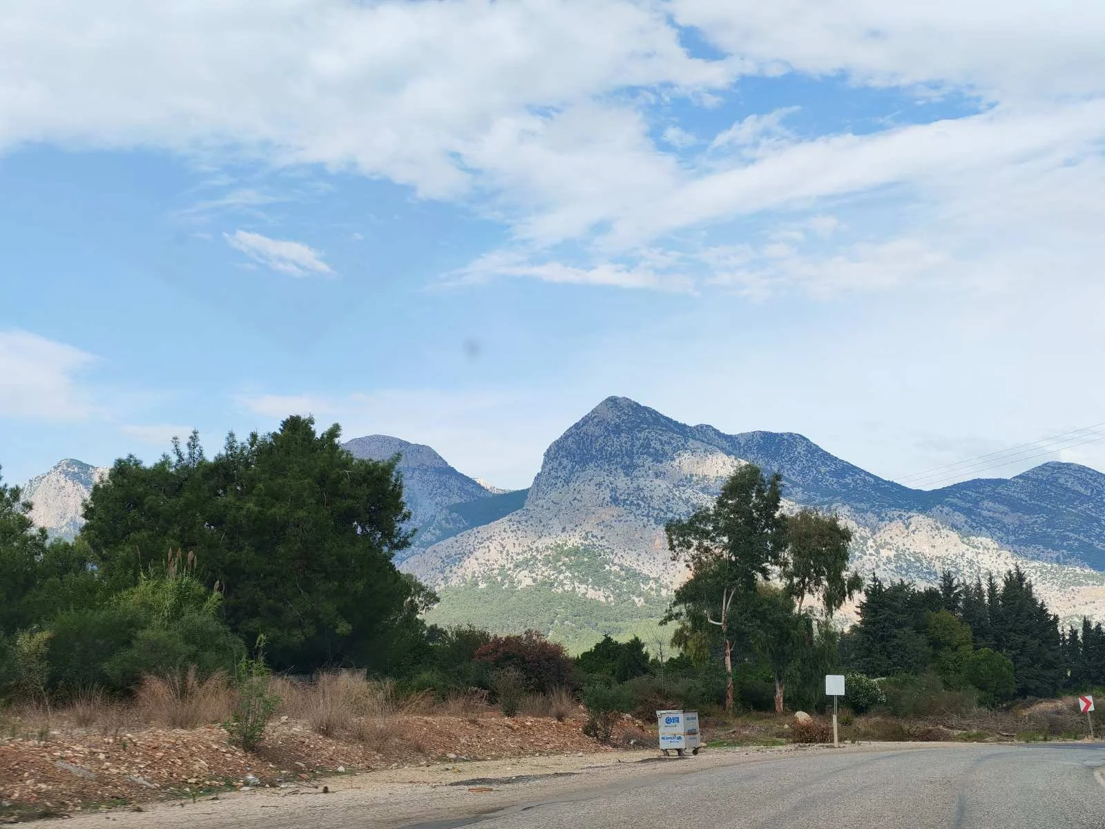 Dağlar ve gökyüzü, ön planda ağaçlar ve yol, bulutlar ve birkaç araç.