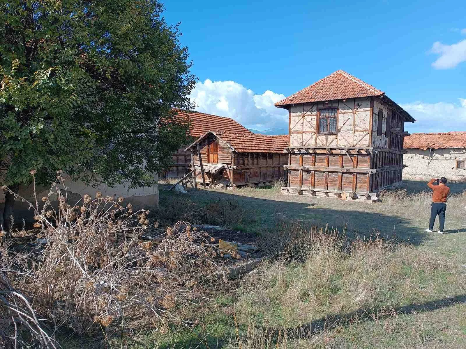 Ağaç ve kuru çiçeklerle çevrili, tahta ve taş yapılı eski köy evi ve bir kişi yanındaki alan.