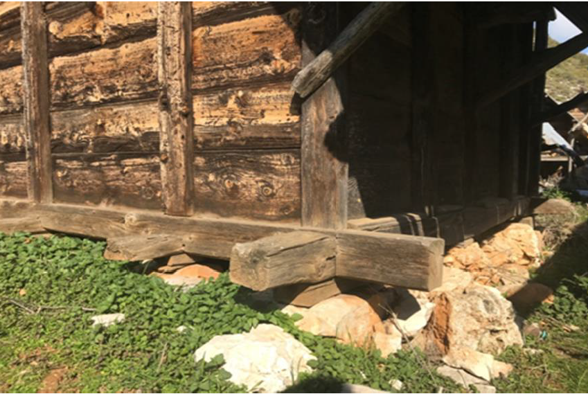 Ahşap bir evin alt kısmı, temel taşları ve yeşil çim ile çevrili.