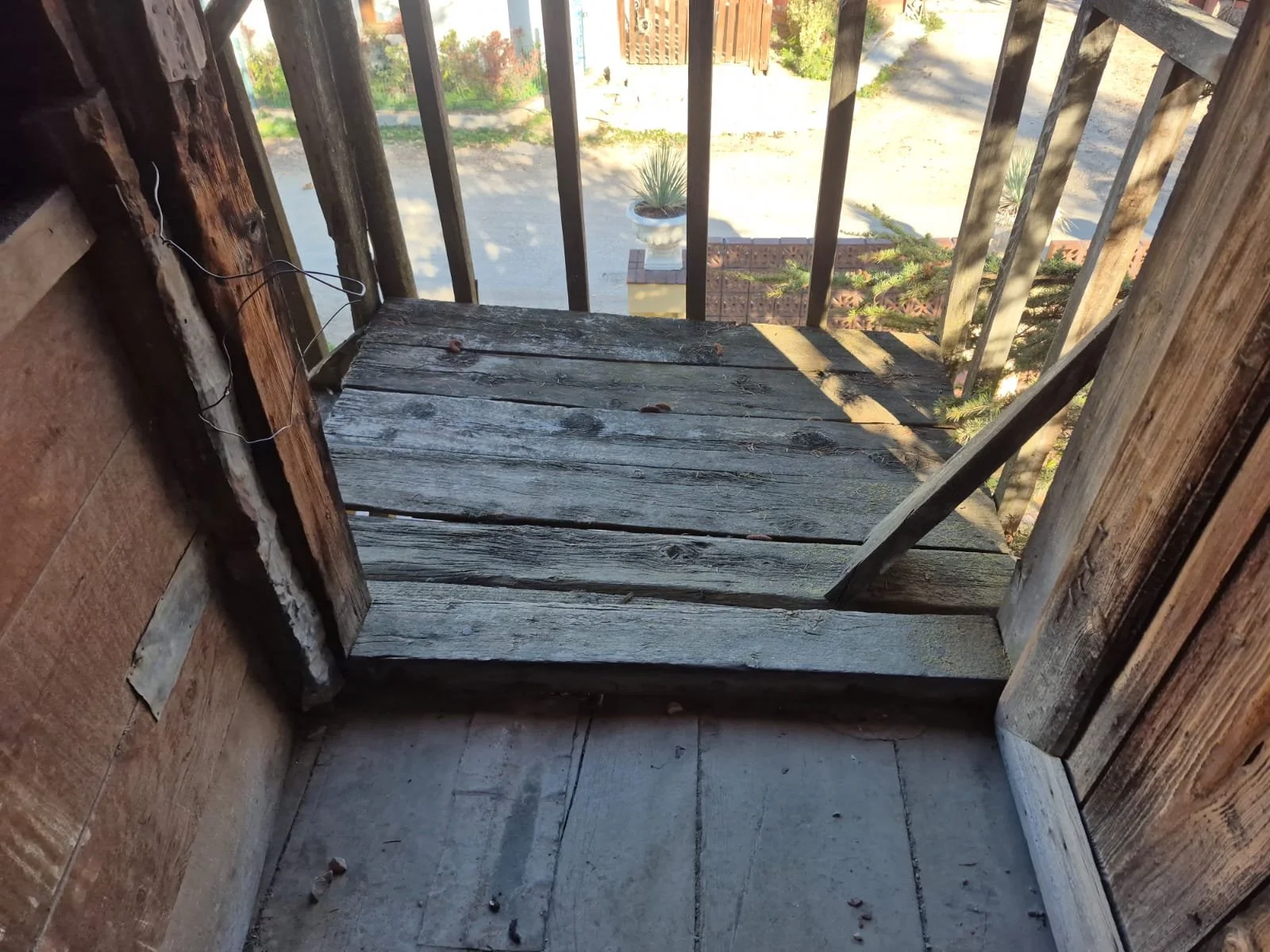 Ahşaptan yapılmış küçük bir balkon veya teras, üstü açık, içeride ve dışarda gölgeler ve güneş ışınları var.