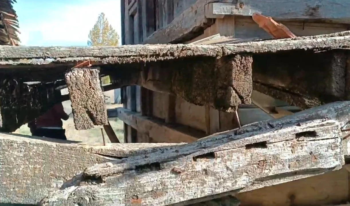 Eski ahşap ve taş malzemeden yapılmış bir iskele veya platform, zamanla aşınmış ve hasar görmüş. Arka planda ise bir yapı ve birkaç ağaç görülüyor.