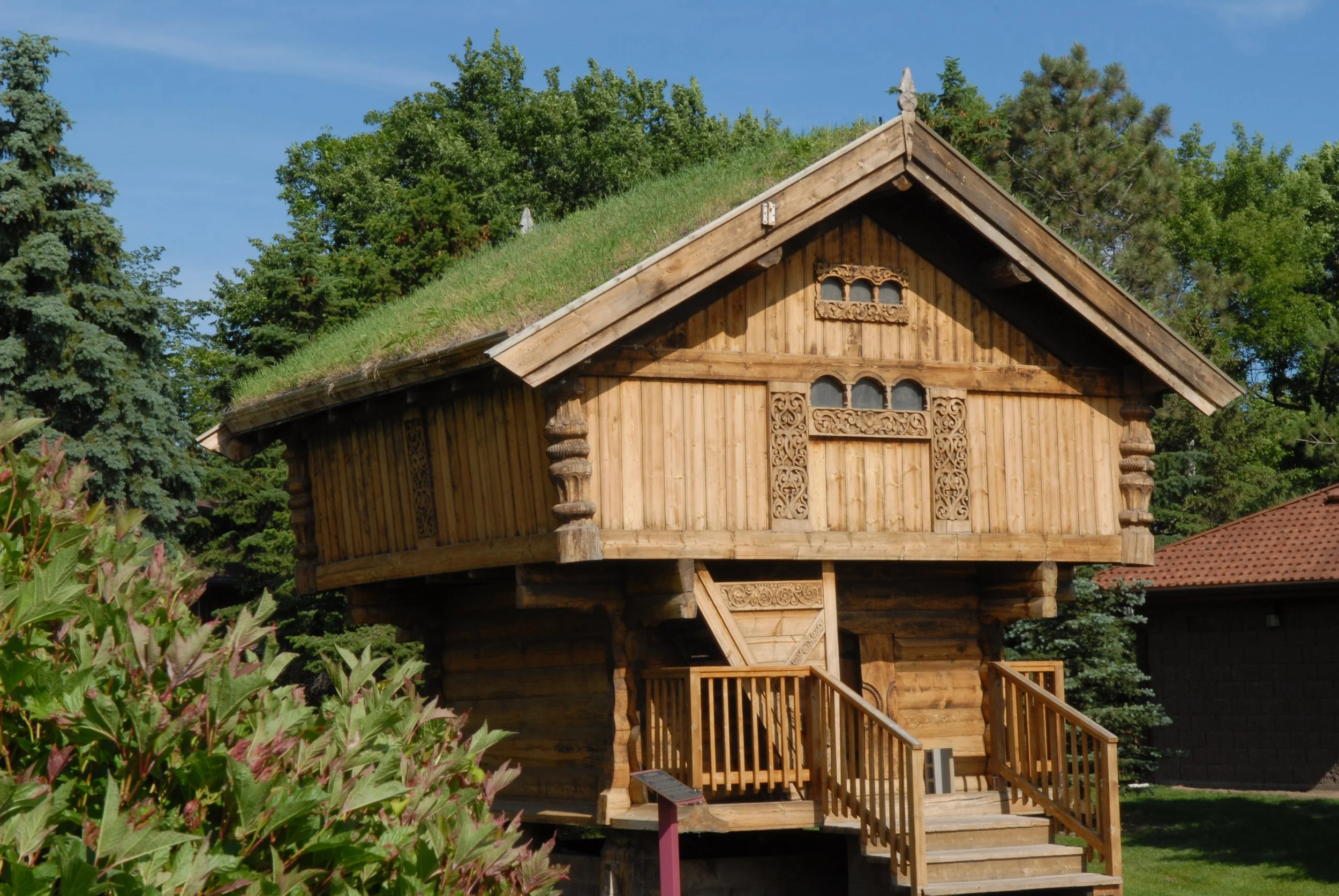 Ahşap yöresel ev, ahşap oymalarla süslenmiş, bahçede, ağaçlar ve bir başka bina arka planda, gökyüzü açık ve temiz.