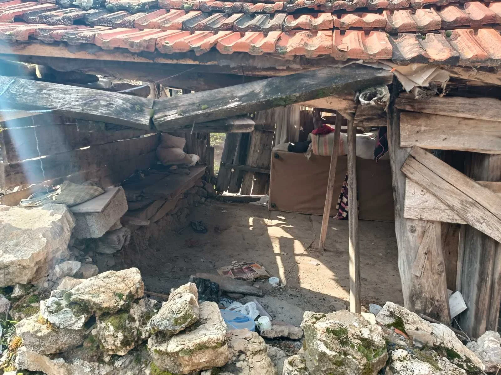 Ahşap ve taş malzemelerden yapılmış, hasar görmüş küçük ve eski bir baraka veya kulübe. Çatısı kırık ve düzgün görünmüyor. İçerisinde bazı eşyalar ve yığılmış odunlar bulunuyor. Dış çevresi taş duvarlarla çevrili. Güneş ışığı içeride hafifçe gölgeler