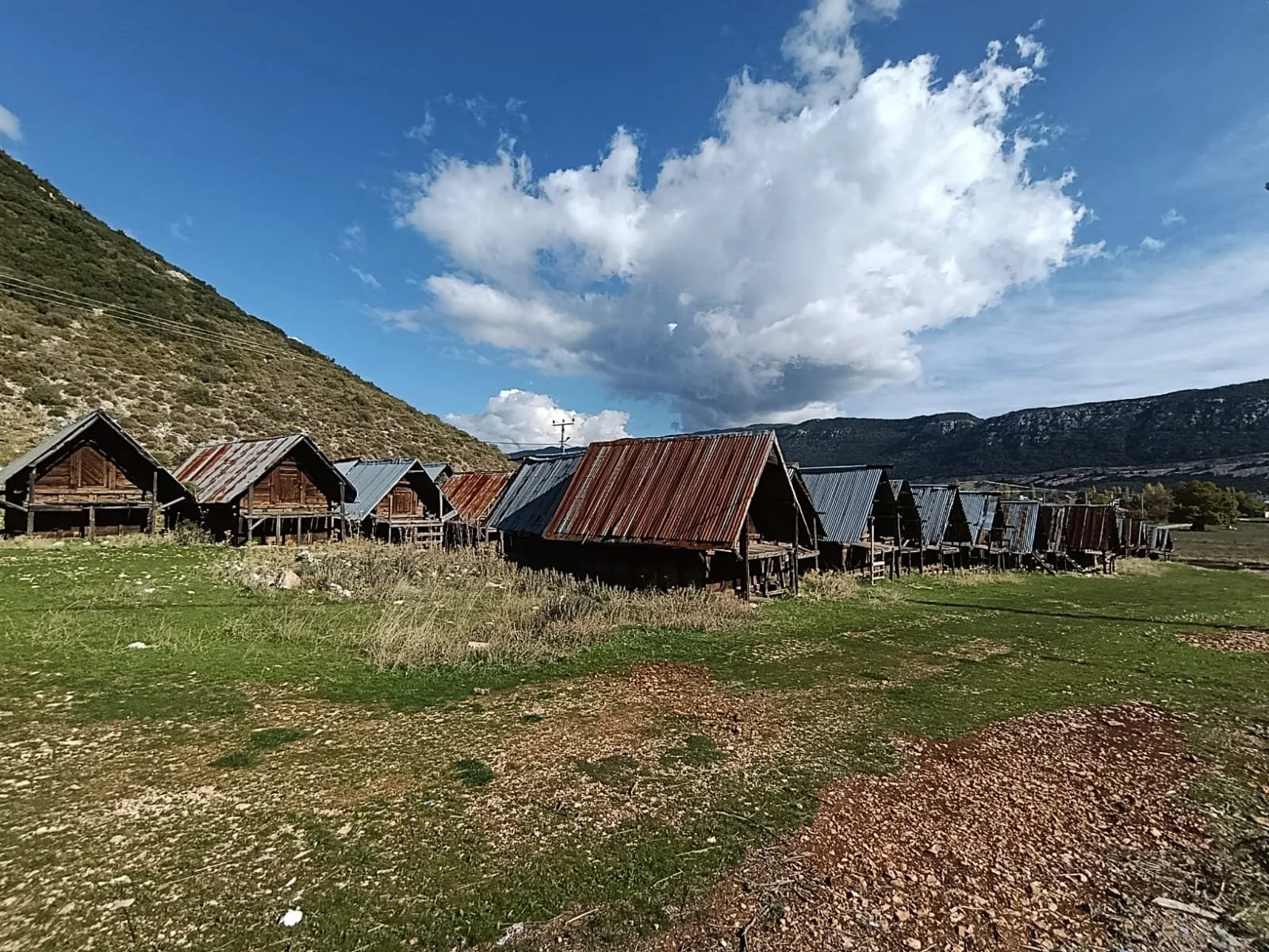 Dağ eteklerine inşa edilmiş birçok ahşap kulübe, gökyüzü ve bulutlar altında yer alıyor.