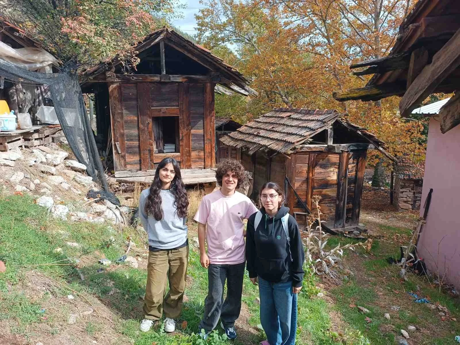 Üç genç kadın, çatlak ve eski ahşap evlerin bulunduğu kırsal bir bölgede duruyor, arka planda ağaçlar ve sonbahar yaprakları görülüyor.