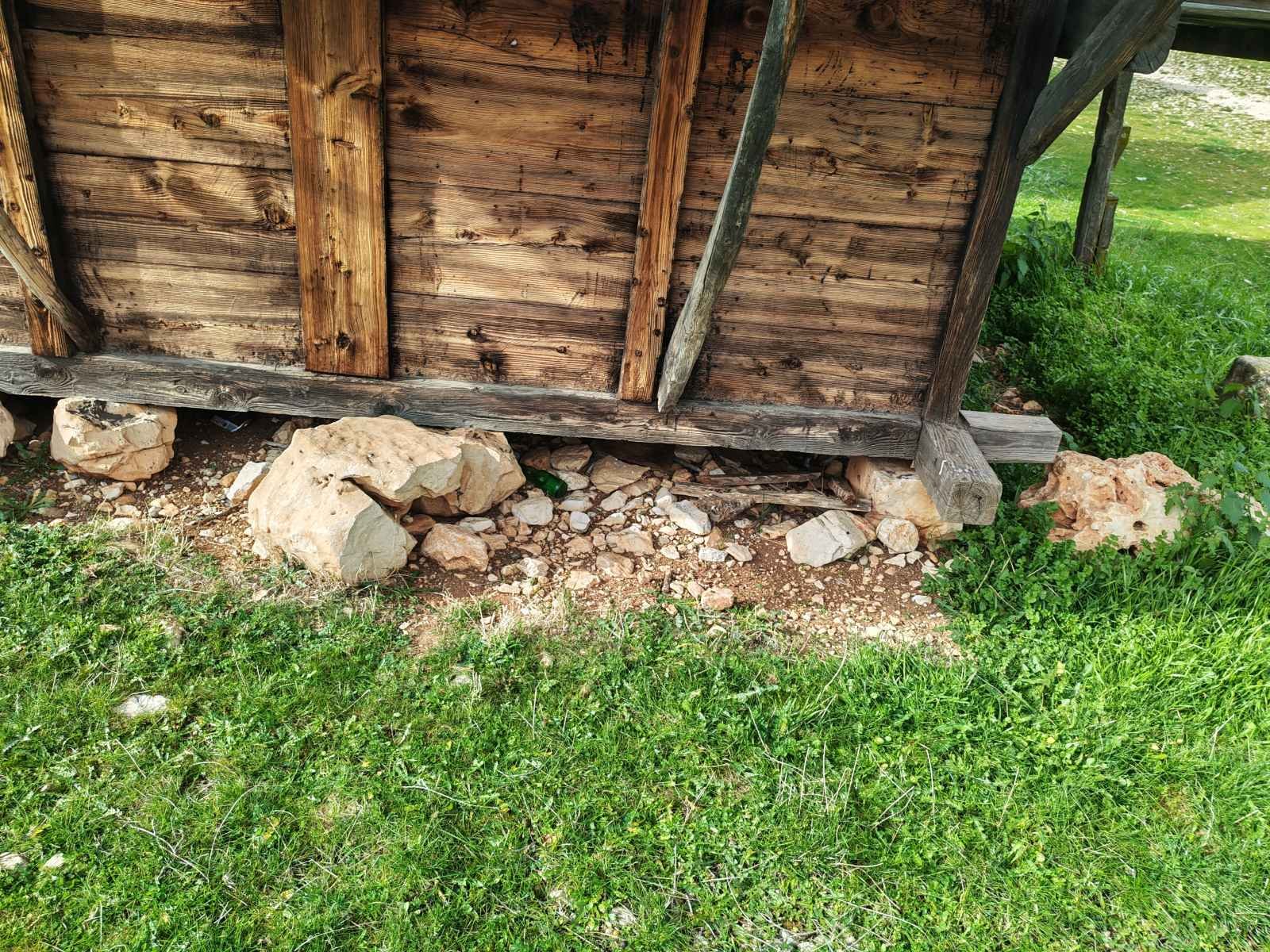 Ahşap bir yapının temel kısmı, taşlar ve toprak üzerinde duruyor, çevresinde yeşil otlar var.